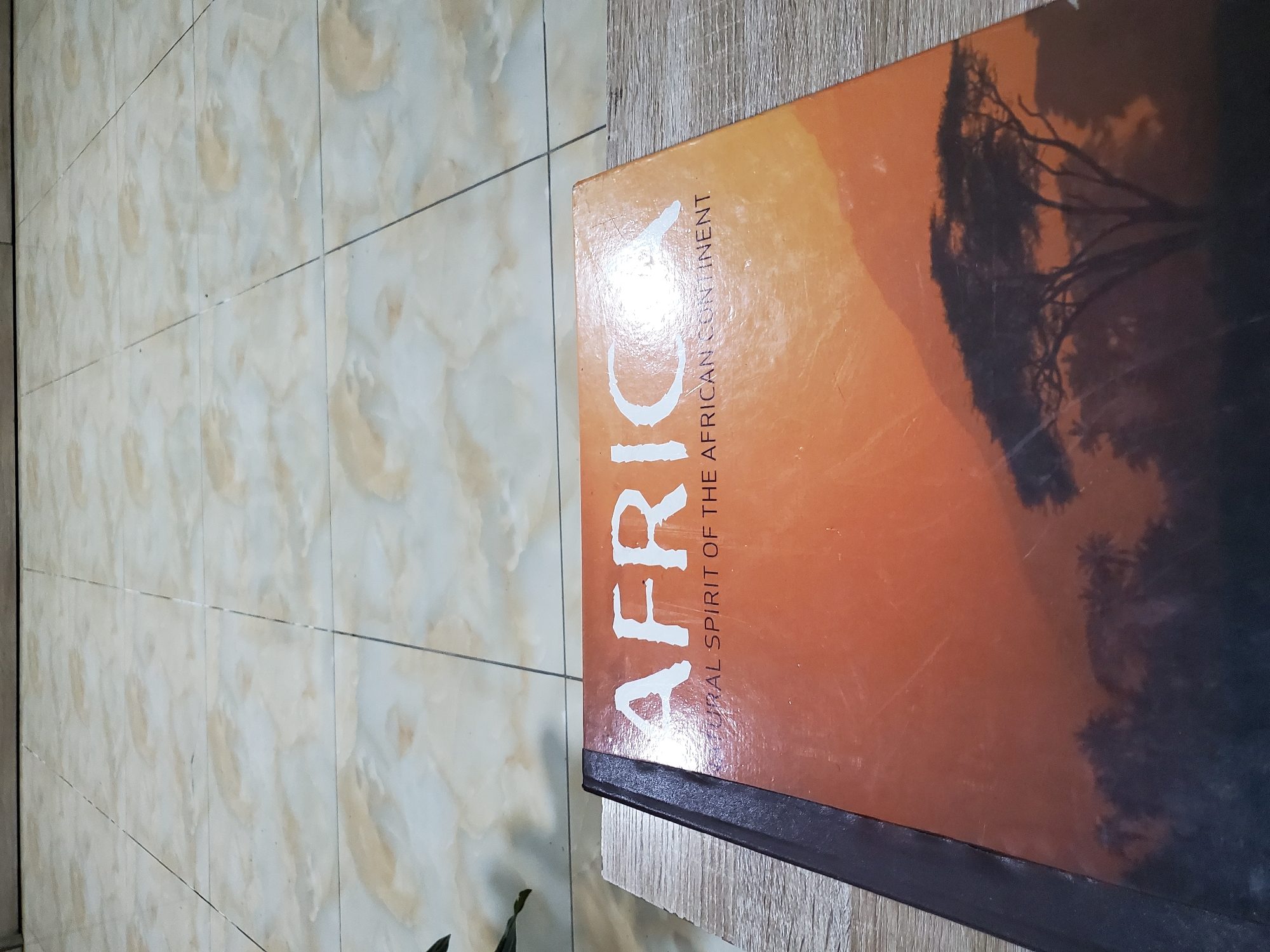 A close-up shot focuses on the cover of a book titled "AFRICA CULTURAL SPIRIT OF THE AFRICAN CONTINENT." The book has an orange cover with a silhouette of an acacia tree against a sunset. The text is in white, with "AFRICA" prominently displayed at the top. The book is placed on a wooden surface. The background shows a tiled floor. There are no people or actions depicted in the image. The location in Jos, Nigeria cannot be determined from the image.