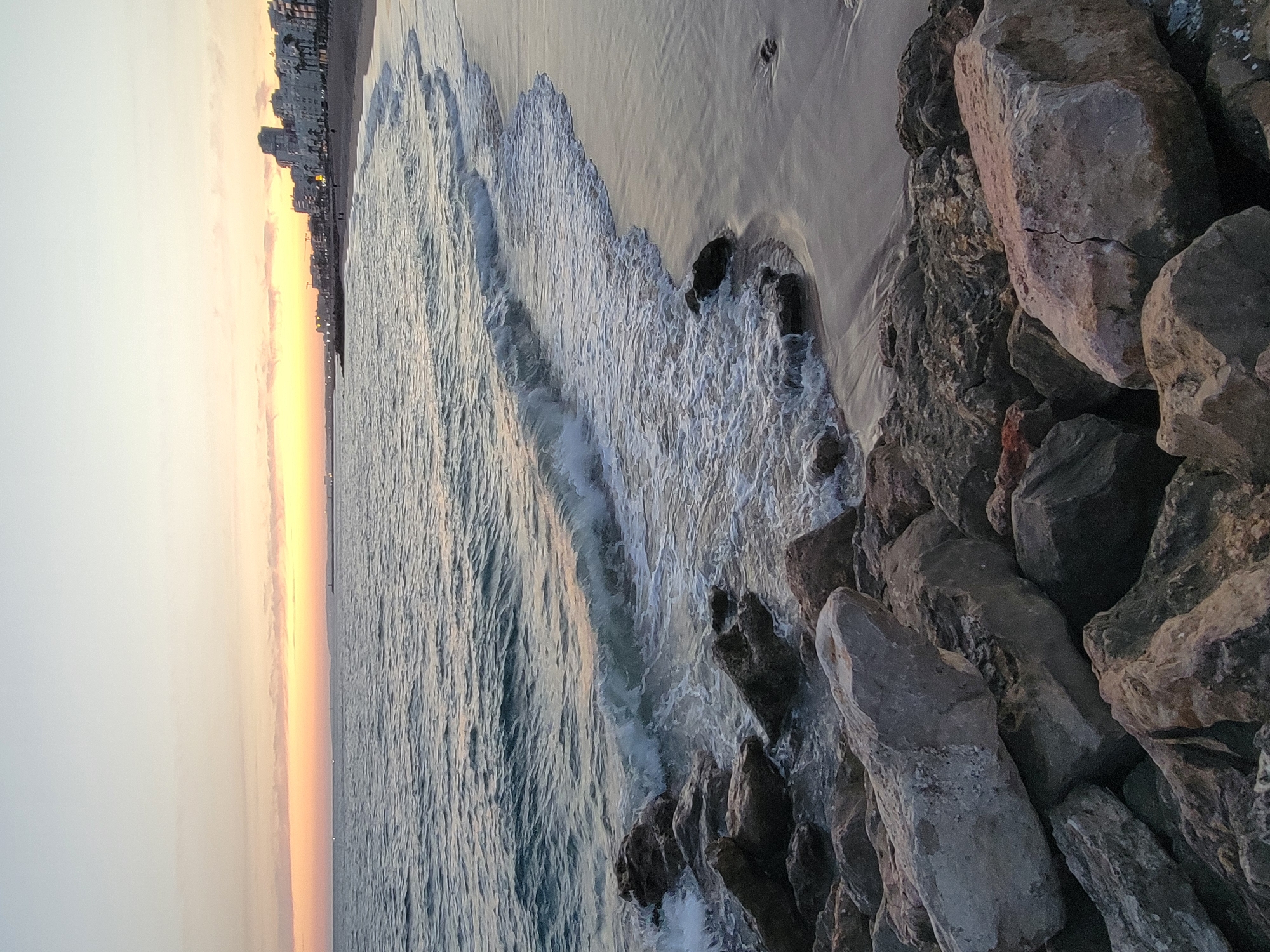 The image depicts a serene beach scene in Quarteira, Portugal, at dusk. The sky displays a gradient of soft pastel colors, transitioning from a pale yellow near the horizon to a light grey at the top, with a thin band of orange hues suggesting the setting sun. The sea is calm, with gentle waves lapping onto the sandy shore.
In the foreground, a rocky embankment is visible, with large, greyish boulders of various sizes scattered along the edge of the water. These rocks are wet from the sea spray, and some are partially submerged. The sand on the beach appears damp and smooth, reflecting the soft light of the sky.
Further down the coastline, a promenade or walkway runs parallel to the beach, lined with apartment buildings and hotels. These structures are silhouetted against the twilight sky, with some lights visible in the windows. A few indistinct figures can be seen walking along the beach and the promenade, suggesting a quiet evening atmosphere. The overall impression is one of tranquility and the peaceful end of a day at the seaside.