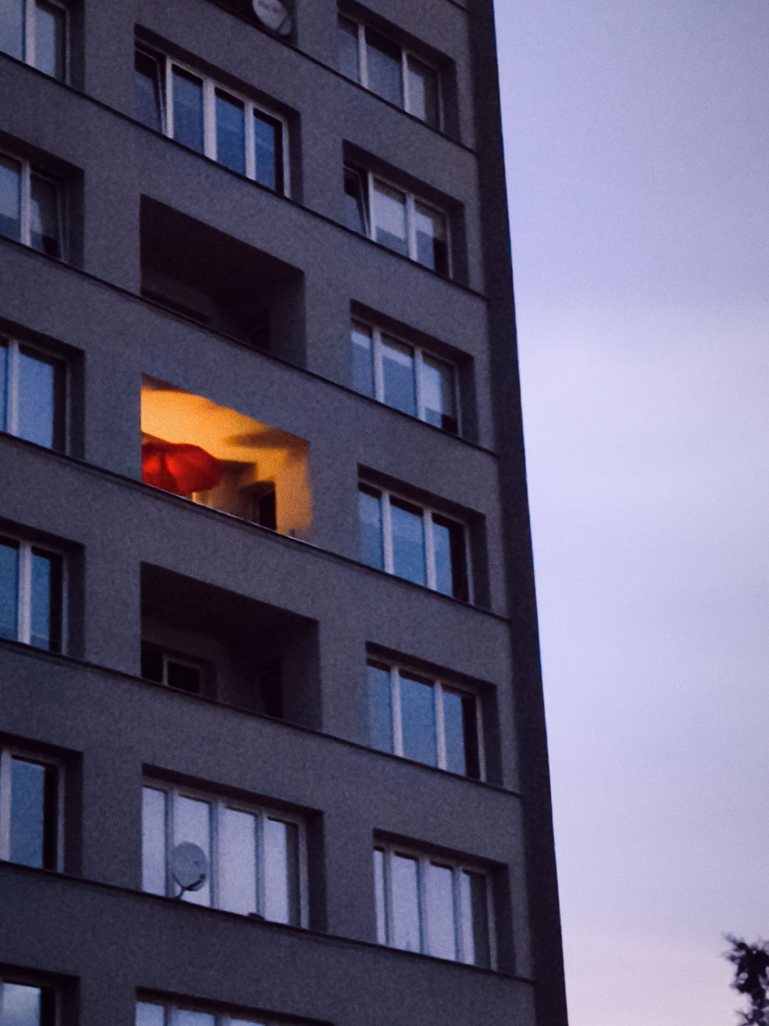 A medium shot captures the side of a dark gray, multi-story apartment building against a pale purple sky. The building features numerous windows, many of which are dark or reflect the muted sky. On the fourth floor from the top, one recessed balcony is illuminated with a warm orange glow, revealing a large, red, billowy object, possibly a piece of fabric or a large balloon. Another window on the seventh floor from the top has a white satellite dish mounted inside. The lower floors of the building also have windows, with the third floor from the bottom showing reflections of the sky. The image appears to be taken in Gdańsk, Poland, likely in the late evening or early morning.