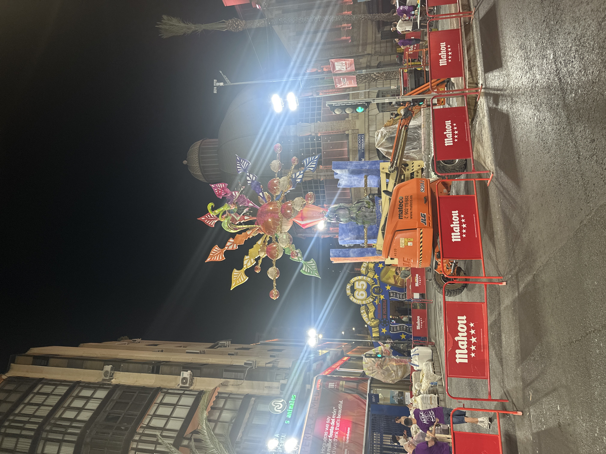 The image depicts a nighttime street scene in Alicante, Spain, possibly during a festival or event. A large, colorful, illuminated decoration resembling a sunburst with leaves hangs in the foreground, casting a vibrant glow. In the background, a building with a dome-like structure is visible, illuminated by bright lights.
Several people are present, some gathered near barriers, while others appear to be working on a scissor lift in the middle of the street. The street is lined with red metal barriers, many of which display the "Mahou" logo with stars. There's also a visible sign for "PESCADOS Y VERDURAS," indicating a market or food stall. The presence of a scissor lift and barriers suggests some form of setup or maintenance for an event. The street appears wet, possibly from recent rain or cleaning. The overall mood seems festive, given the decorations and lights, but also functional with the presence of work equipment. Text visible includes brand names like "Mahou," "mateco," and "JLG," as well as the Spanish phrase "Hogueras we say" and "millor festa del món," which translates to "best festival in the world," strongly suggesting the event is related to the "Hogueras de San Juan" festival in Alicante.