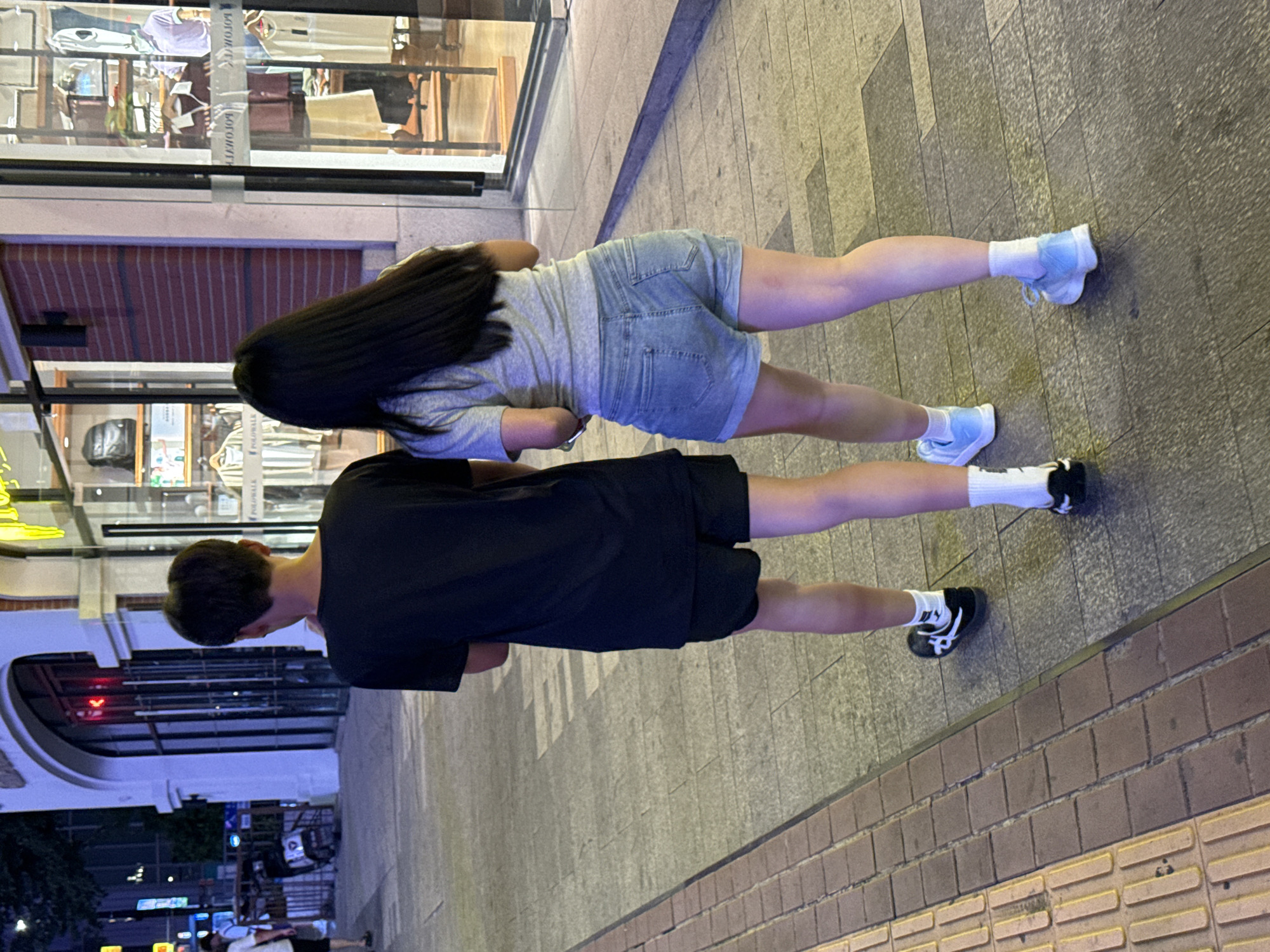 In Yuzhong District, China, a young man and woman walk side-by-side on a paved sidewalk at night. The man, seen from behind, wears a black t-shirt and black shorts. The woman, also seen from behind, wears a grey t-shirt and denim shorts. They are walking away from the camera, towards a brightly lit store with large windows displaying clothing and mannequins. The sidewalk is a mix of grey concrete and brick. Streetlights illuminate the scene, casting shadows on the ground. In the background, building facades and a parked scooter are visible.