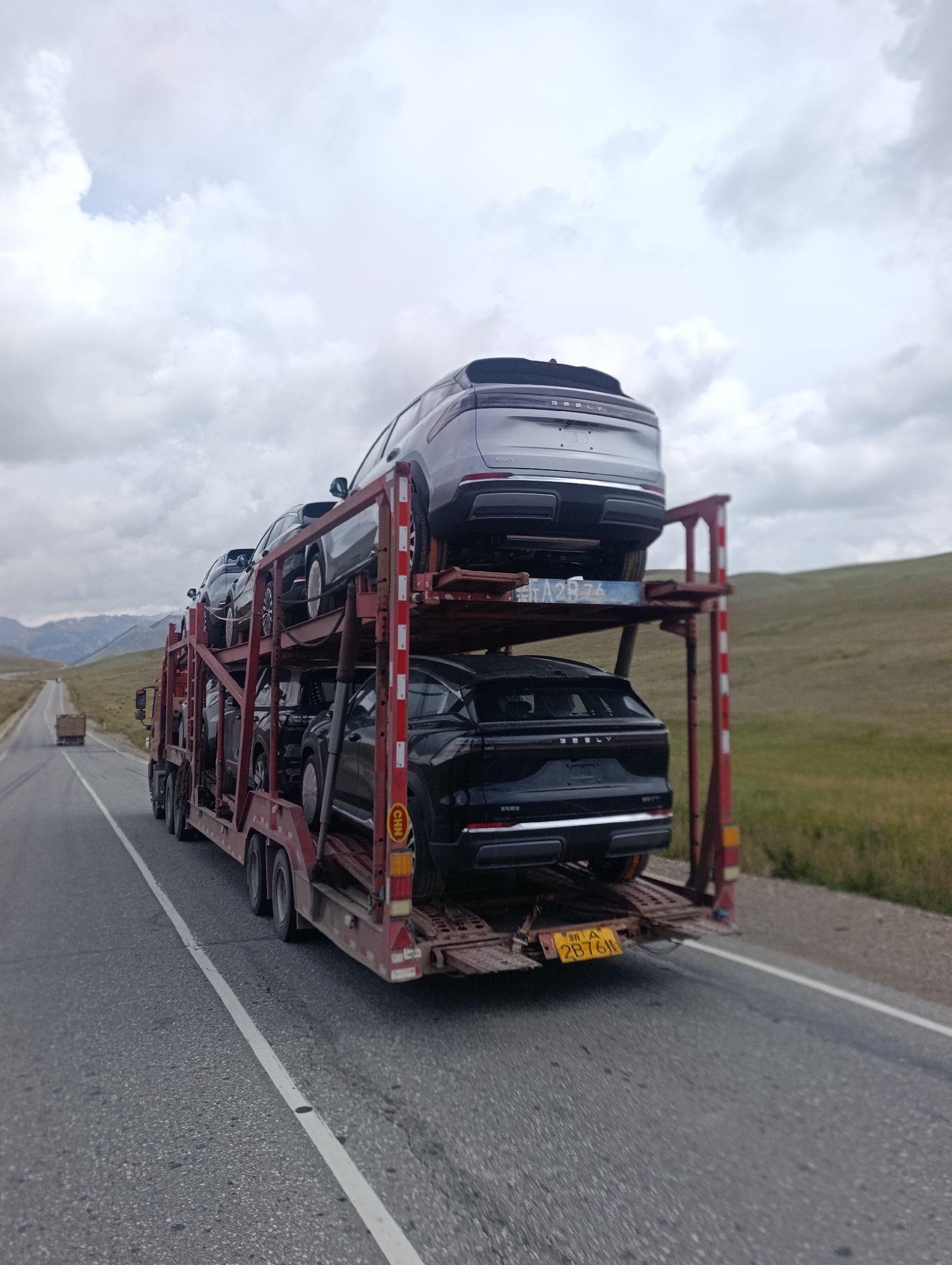 A car carrier truck is driving on a two-lane asphalt road in a rural, mountainous region. The truck is loaded with several new, dark-colored SUVs. The sky is overcast. The landscape on either side of the road consists of rolling green hills and distant mountains. The road appears to be in Kyrgyzstan, likely near the city of Ак Жар.
