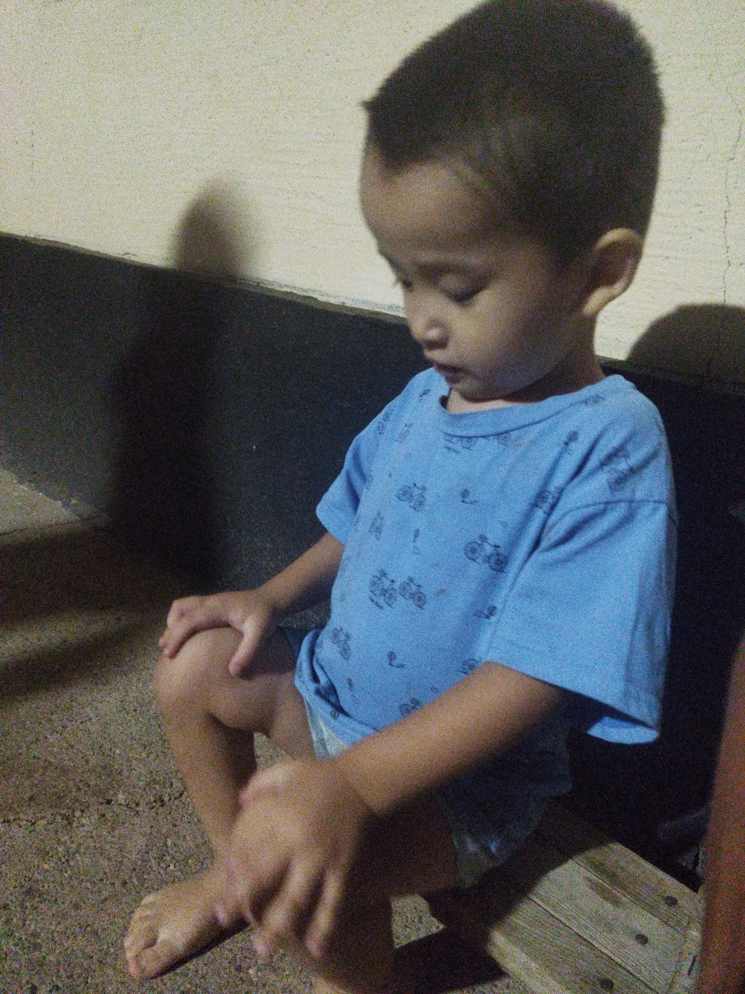 A young child, appearing to be of Asian descent, sits cross-legged on a rough, sandy ground in front of a wall. The child wears a blue t-shirt with a pattern of bicycles and is looking downwards, with their hands resting on their knees. A shadow is cast on the wall behind them, suggesting artificial lighting. To the right of the child, a portion of a wooden surface is visible, possibly a bench or stool. The overall environment appears to be an outdoor setting, possibly an alleyway or a less-maintained area. The image exhibits significant grain and noise. The location is Mailuu-Suu City, Kyrgyzstan.