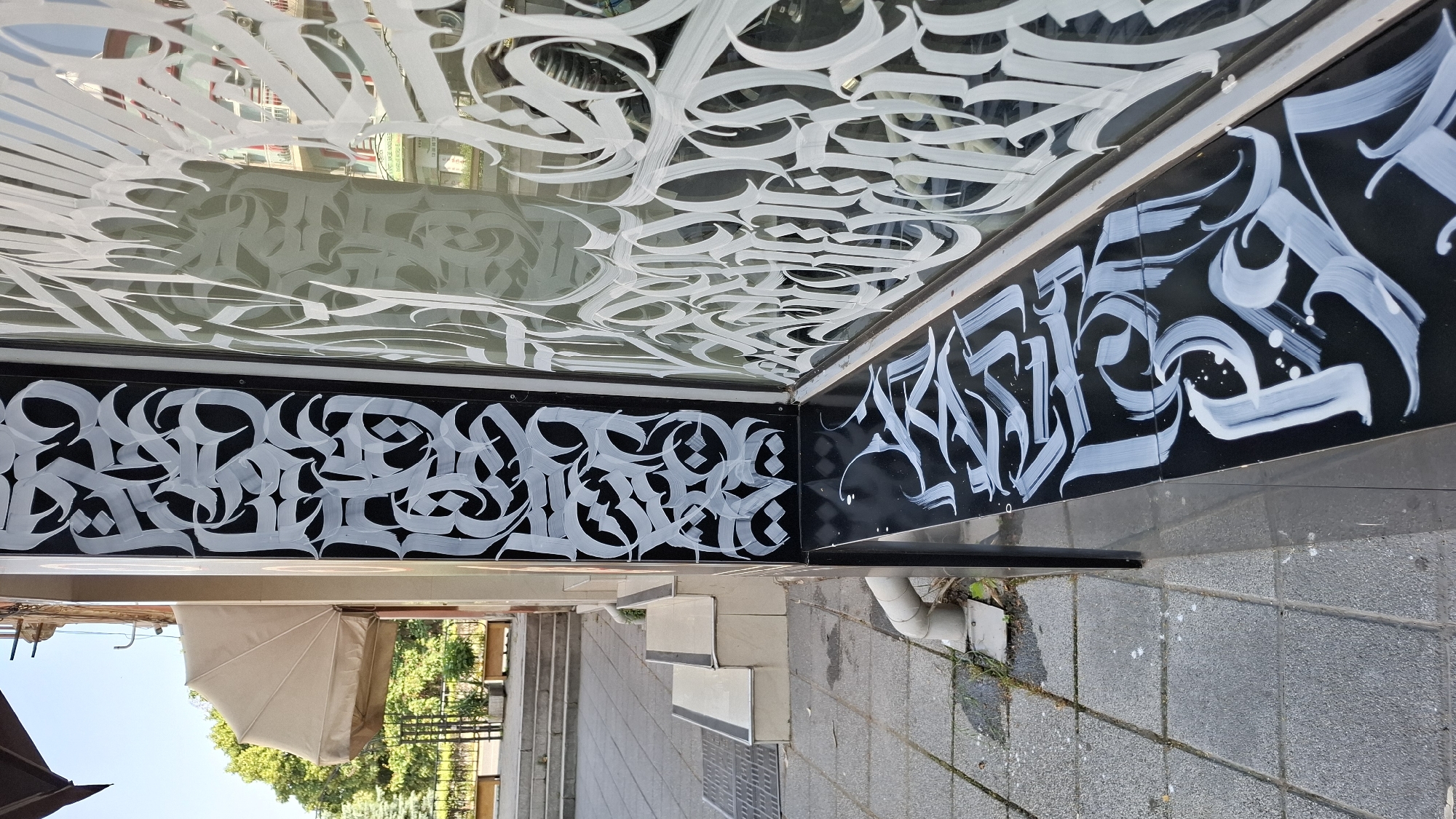 The image shows a section of a building facade in the city of Plovdiv, Bulgaria. On the left, a tan awning is partially visible above a set of concrete stairs. The main focus of the image is the black surface of the building, which is adorned with intricate white calligraphy. This calligraphy appears on both a vertical section to the left and a horizontal ledge extending to the right. The calligraphy is stylized and decorative, with flowing lines and sharp angles. A large glass window to the right also features white calligraphy, which appears to be the same design as on the ledge, creating a reflective and layered effect. The ground in front of the building is paved with grey square tiles. A white downspout is visible on the left side of the image, near the tiled ground. The scene appears to be outdoors on a sunny day. There are no people visible in the image.