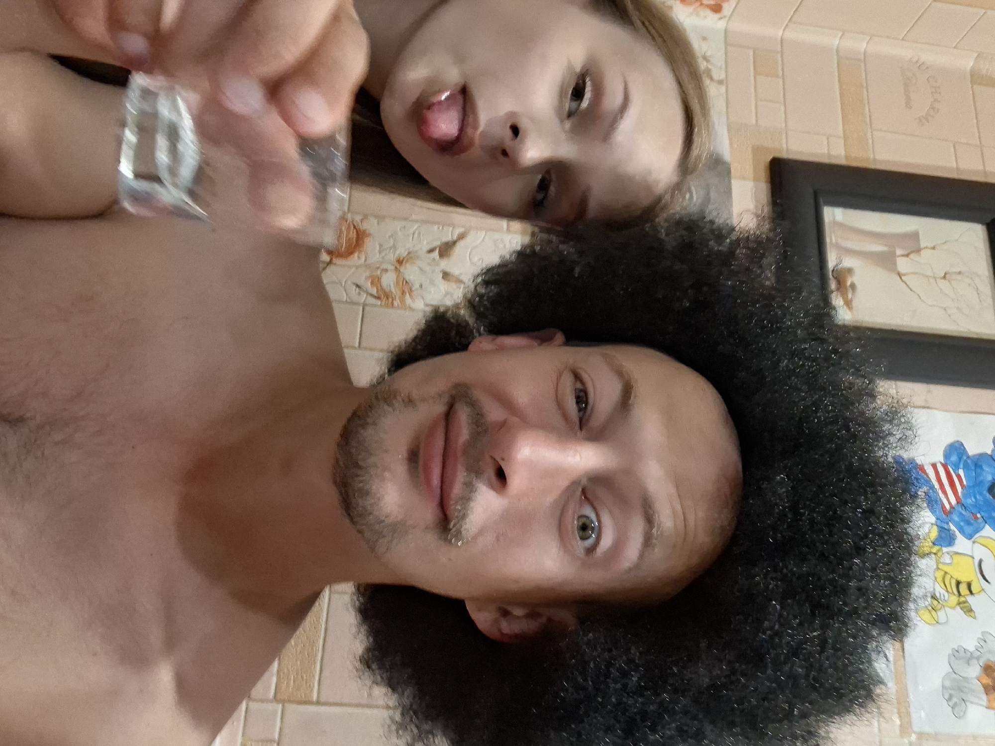 Two individuals, a male and a female, are captured in a close-up selfie, likely in a bathroom given the tiled walls and a framed picture. The male, positioned in the foreground and to the right, sports a large afro wig and has a shaved head with a goatee and mustache. His left eye is wide open, while his right eye is partially closed, giving a peculiar expression. He is shirtless. The female, to the left and slightly behind, has blonde hair and is sticking her tongue out, holding up a small, clear shot glass filled with a clear liquid. She is also shirtless. The background includes beige tiled walls with a subtle floral pattern, and a framed picture on the wall depicting a still life with vases and branches. There are also some cartoon drawings partially visible at the top. The overall scene appears to be informal and possibly playful. The location is not discernible from the image itself, and no information about Myadzyel, Belarus is present.