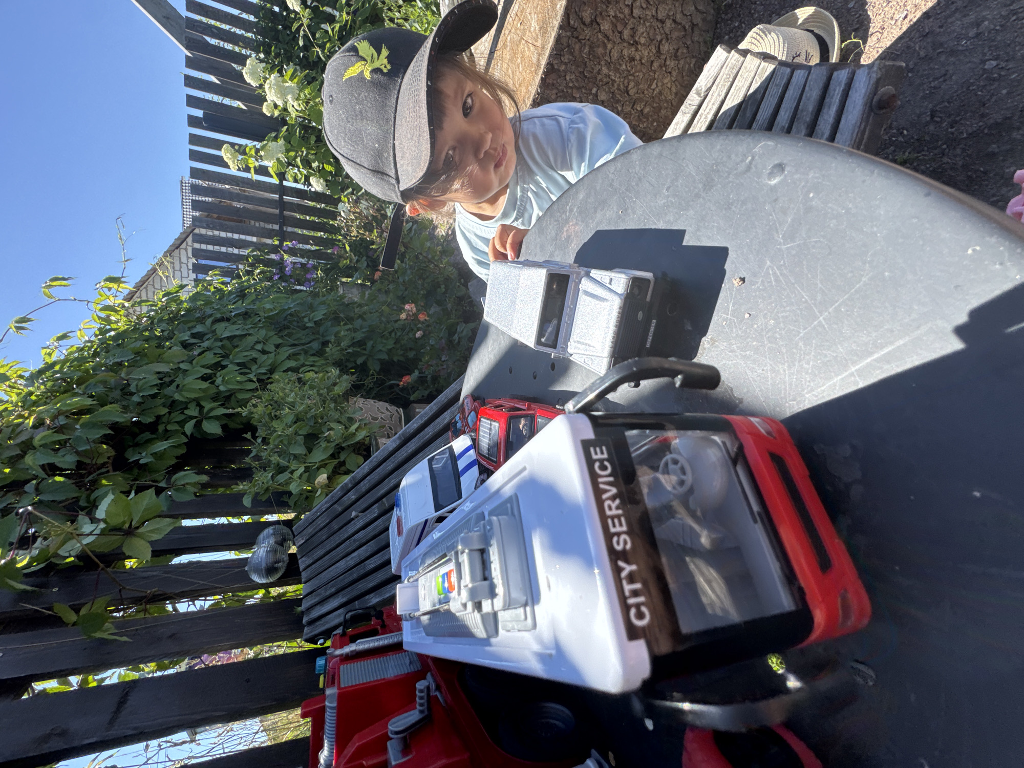 In Kempelevo, Russia, a young child wearing a black baseball cap and a light blue t-shirt is seated at a round, gray table outdoors. The child is leaning forward, looking at the camera with a pensive expression. In front of the child on the table are several toy vehicles: a silver SUV, a white bus with "CITY SERVICE" written on it, and a red car. A red toy fire truck is partially visible on the left side of the frame. The background features a wooden fence, lush green foliage, and flowering plants. The sky is clear and blue. A straw hat rests on a wooden bench to the right of the table.