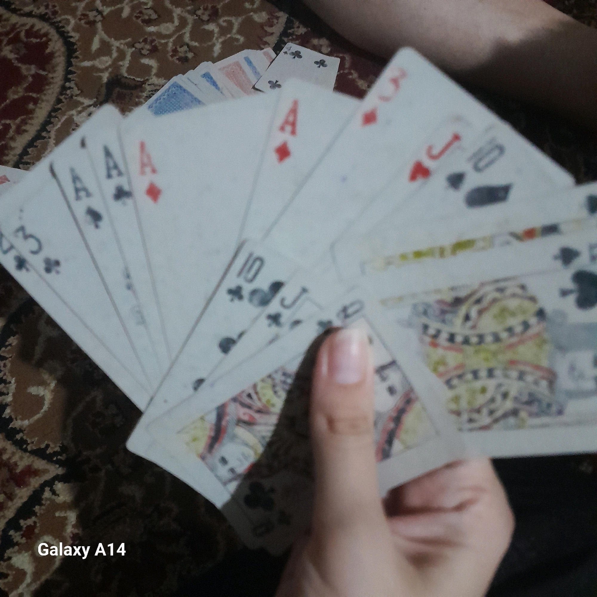 A close-up, slightly blurry, overhead shot shows a fan of playing cards held by a person's hand. The cards are fanned out in a semicircle on a patterned rug. The visible cards include Ace of Spades, Ace of Hearts, 3 of Clubs, Ace of Diamonds, 3 of Diamonds, 10 of Clubs, Jack of Clubs, and a Queen or King. The background is a dark, patterned rug, possibly from Khan Yunis, Palestinian Territories. The lighting is moderate, casting soft shadows. The "Galaxy A14" watermark is visible in the bottom left corner.