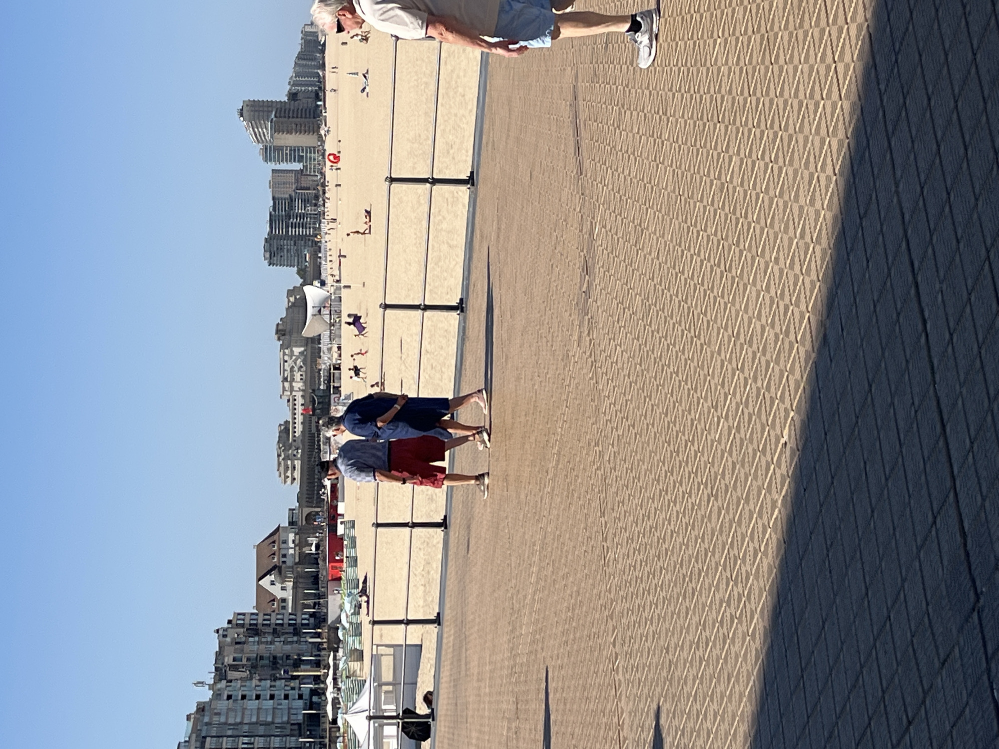 In Ostend, Belgium, on a bright, sunny day, a beach scene unfolds along the coast. The foreground features a textured, tan-colored promenade with a diagonal shadow stretching across it. In the mid-ground, two men, one in a blue shirt and red shorts, the other in a dark blue shirt and shorts, walk along the promenade, facing away from the camera. Behind them, a black metal railing separates the promenade from a sandy beach dotted with numerous beach umbrellas and lounge chairs. Further in the background, several people are scattered across the beach, some sunbathing and others walking. Beyond the beach, a cityscape of multi-story buildings with varying architectural styles lines the horizon under a clear blue sky. To the far right, an older man wearing sunglasses, a light-colored polo shirt, and blue shorts walks on the promenade, his back partially to the camera.