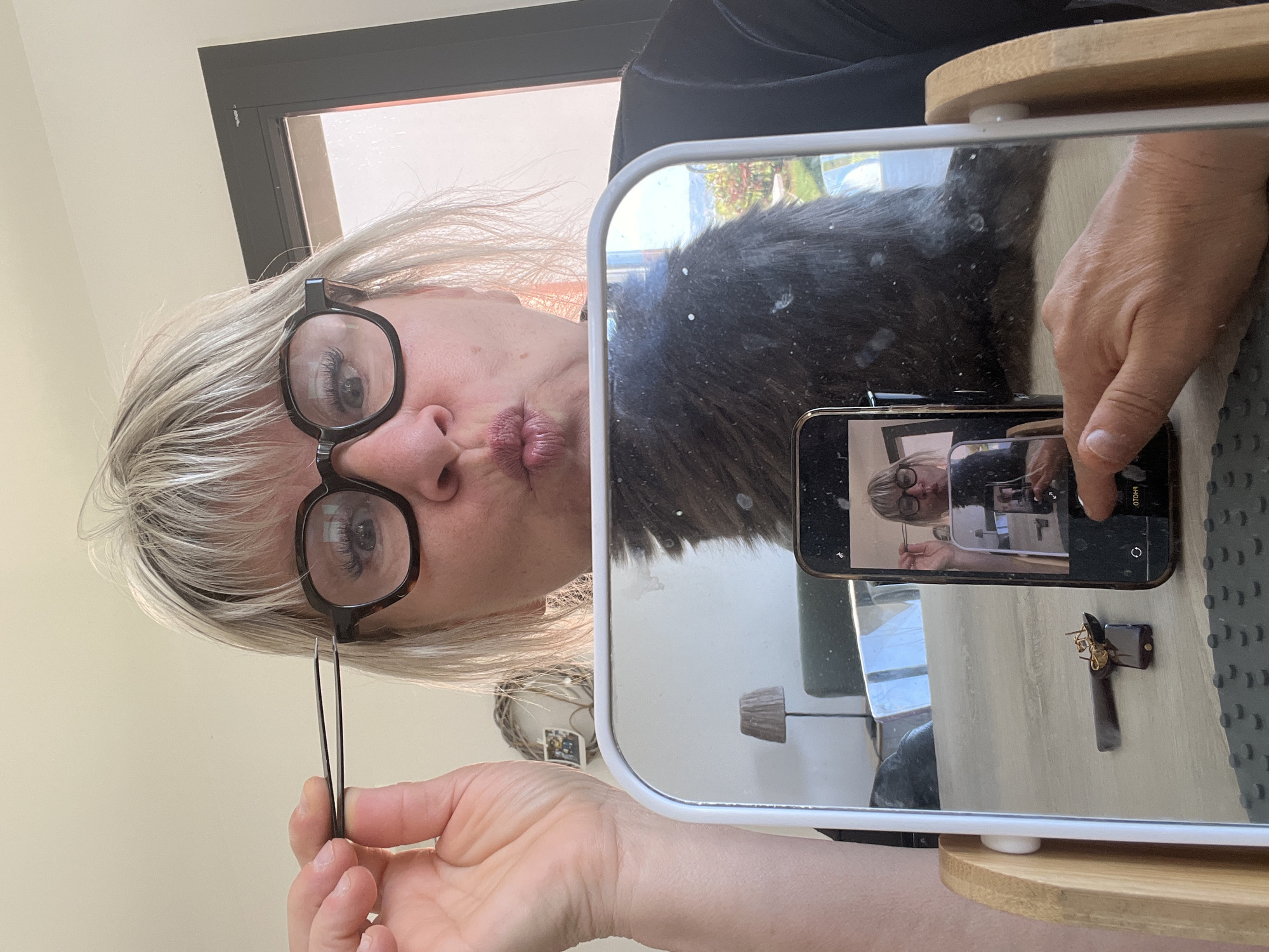 A person with short white hair and glasses is pouting and holding tweezers. A mirror reflects the person and a cell phone taking a photo of the mirror. An object resembling a small beetle-shaped pin lies on a table. The location is Chambœuf, France.