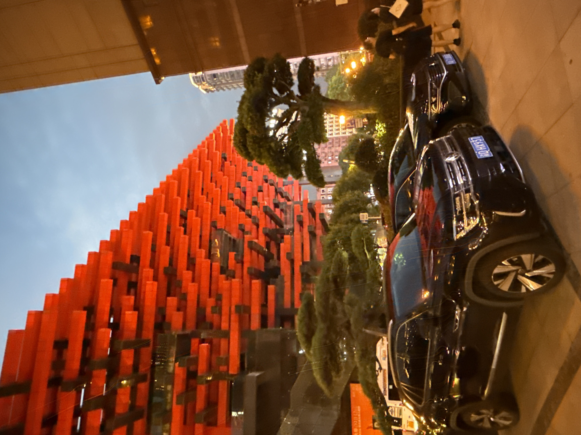 The image shows a street scene in Yuzhong District, China, at dusk. The most prominent feature is a modern building with a facade constructed of numerous bright red, horizontal rectangular elements, layered and staggered to create a striking visual effect against the blue-grey sky. To the right of this building, a tall, tan-colored modern building dominates the right side of the frame. In the foreground, two dark-colored SUVs are parked on a paved area. The SUV on the left is black and appears to be a Volkswagen. The SUV on the right is also dark and partially visible. Several people are visible in the background, including a group of two women dressed in dark clothing standing near the right edge of the image. Behind the parked cars and in front of the red-accented building, there are several well-maintained trees, some appearing to be topiaries or bonsai-style. Streetlights and building lights are illuminated, casting a warm glow on the scene. The overall impression is of an urban environment with distinctive architecture and a lively atmosphere as day transitions into night.