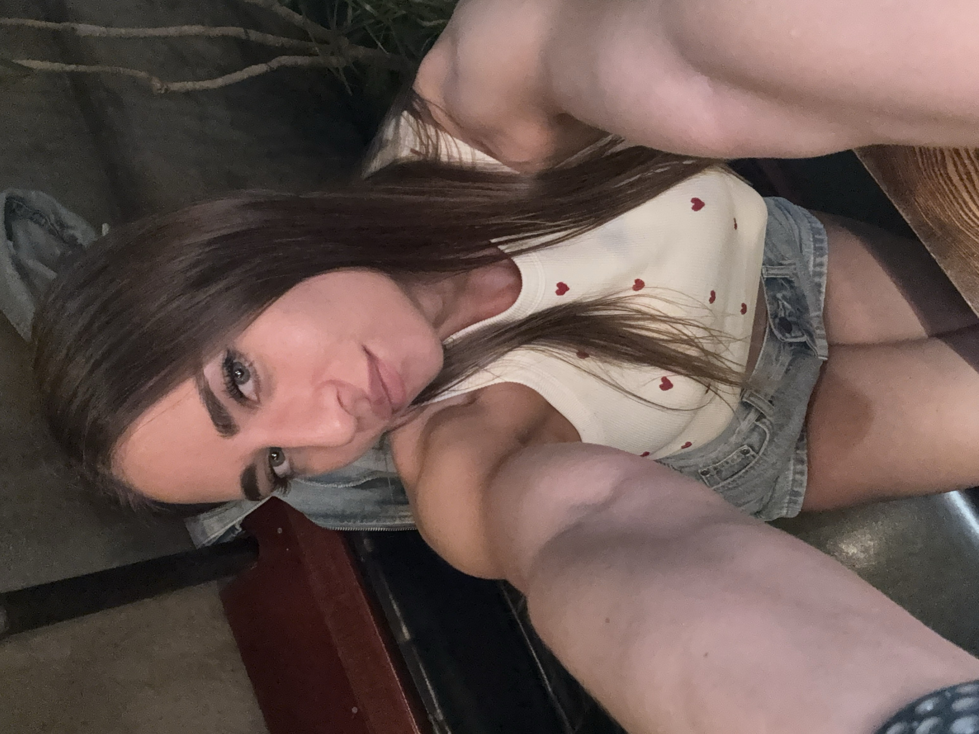 The media file shows a close-up, high-angle, overhead shot of a young woman in Saint Petersburg, Russia. She has long, dark brown hair and is looking towards the camera with a slight smile. Her eyes are open, and she is wearing makeup, including eyeliner and mascara. She is dressed in a white top with small red hearts printed on it and a denim skirt. Her left arm is visible, extended towards the camera, and her right arm is bent and resting on her lap. The background is dark and out of focus, with some indistinct plant-like shapes and what appears to be a wooden surface. The setting appears to be indoors, possibly at a restaurant or bar, with a casual, relaxed atmosphere. The lighting suggests it might be evening or nighttime. The overall emotion conveyed is one of casualness and comfort. There is no visible text in the media file.