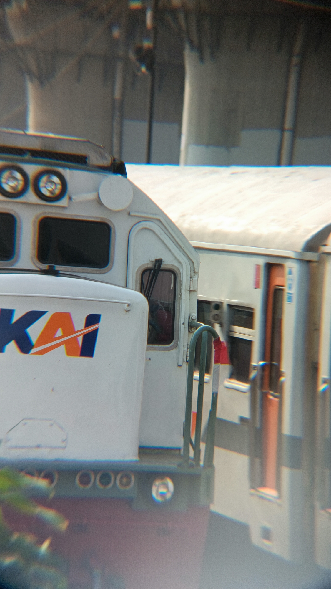 The image captures a close-up, slightly angled view of the front of a white train engine with blue and orange "KAI" lettering. To the right of the engine, another train car is visible, primarily white with an orange door and a dark window. The trains appear to be in motion, evidenced by motion blur and the separation between them. In the background, structures resembling elevated train tracks or an overpass are visible, suggesting an urban or semi-urban environment. The lighting suggests it is daytime, with some sunlight casting highlights on the trains. The foreground shows some out-of-focus green foliage. The image is set in Margajaya, Indonesia, a location known for its railway infrastructure.