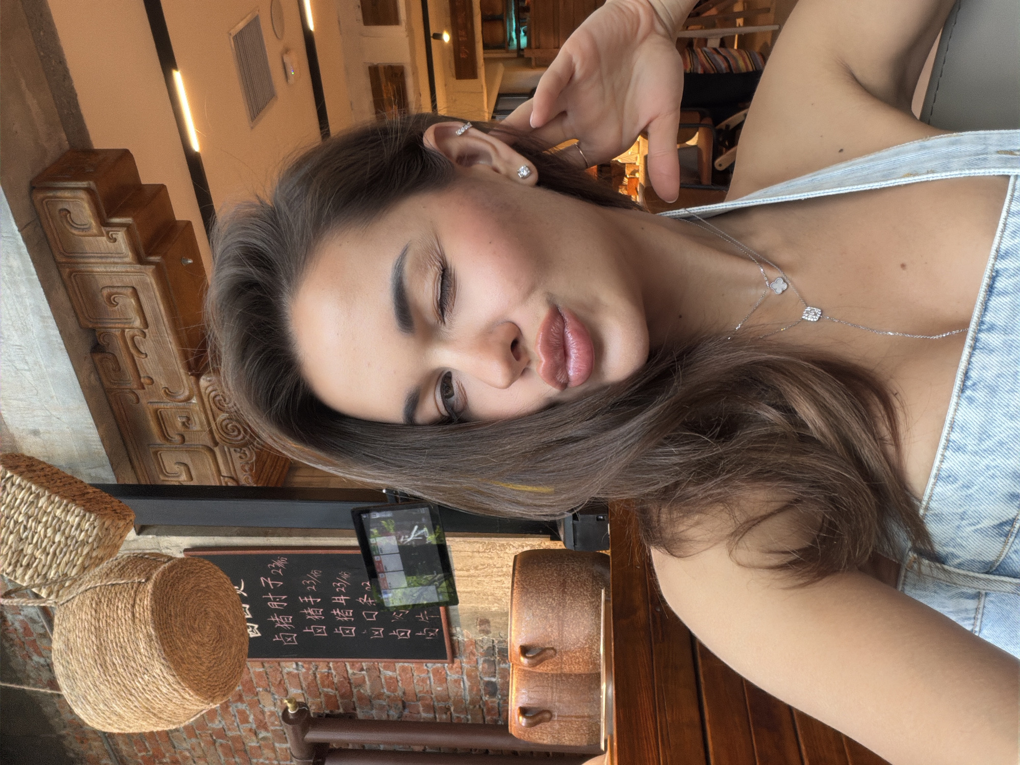 The image is a selfie of a woman with brown hair, wearing a denim top and a silver necklace with a clover and a square pendant. She is winking and puckering her lips, with her left hand raised to her head. She has multiple earrings on her left ear.
The background includes interior elements such as brick walls, a chalkboard menu with Chinese characters (including the price of "卤猪肘子" pork knuckle, "卤猪手" pig trotters, "卤猪耳" pig ears), woven baskets hanging from the ceiling, a television screen, and wooden furniture.
Based on the Chinese characters on the menu, the presence of "卤" (lu, braised) dishes, and the overall aesthetic, the location appears to be a restaurant in a Chinese-speaking area. While I don't have explicit data to definitively pinpoint the exact location, the menu suggests a focus on traditional Chinese cuisine. I cannot confirm that the location is in the Chaoyang District of China.