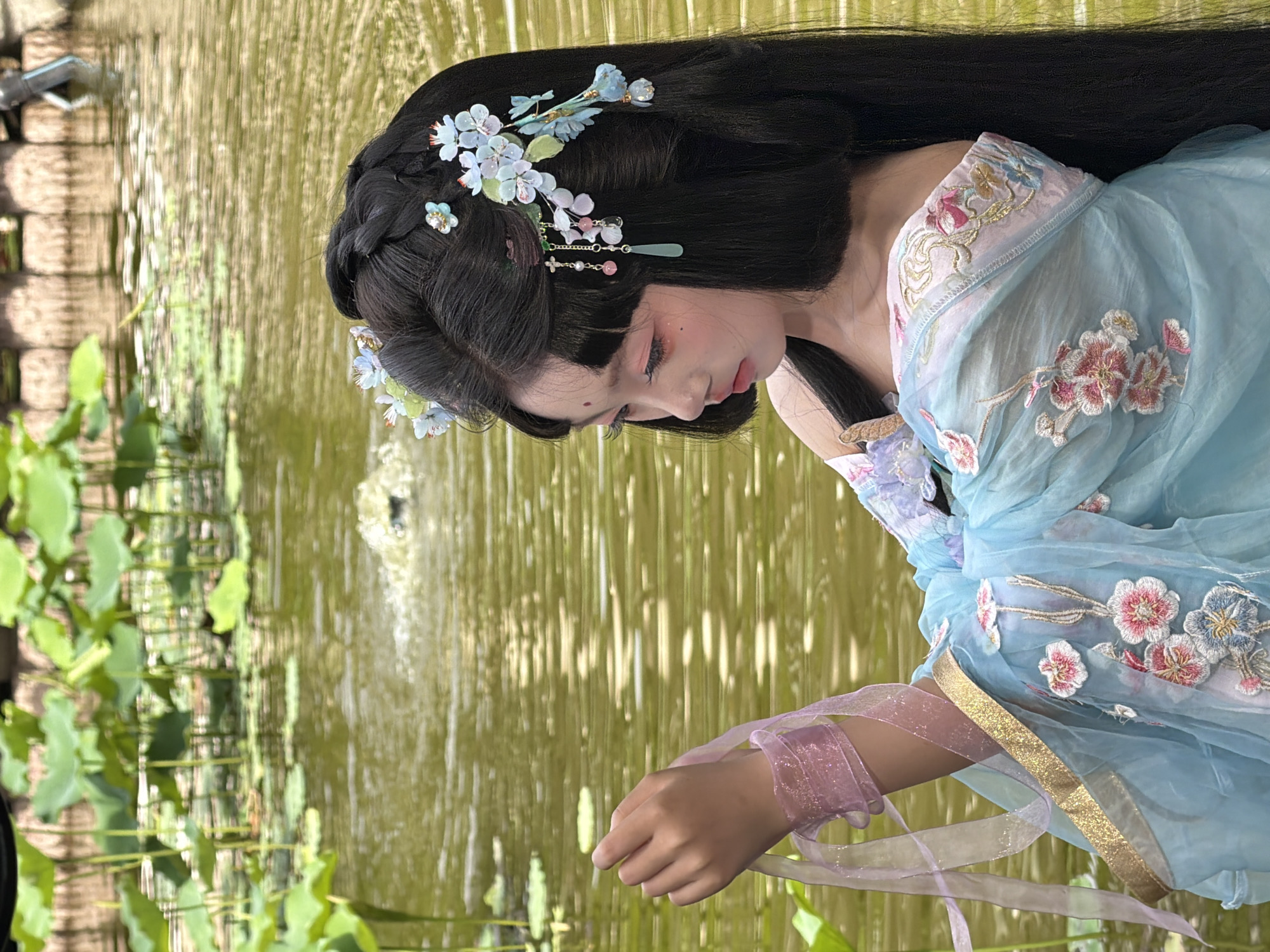 A young East Asian female with long black hair styled with floral hair ornaments and a red dot on her forehead is positioned in the foreground, to the right of the frame. She is wearing a blue and pink traditional Chinese dress adorned with floral embroidery and gold trim. Her left arm is raised, holding a pink ribbon. She is looking down and to her left with a contemplative expression.
The background features a body of water, likely a pond, with lily pads and vegetation growing in and around it. The water is murky green with reflections of the sky and surrounding greenery. In the far background, there is a textured wall or embankment, possibly made of stone or brick. A fountain or water feature is active in the water, creating a small disturbance.
The image appears to have been taken outdoors in a natural setting, possibly a garden or park, near a body of water. While the location is stated to be Yuzhong District, China, this cannot be visually confirmed from the image alone. The lighting suggests it is daytime, with soft, diffused light.