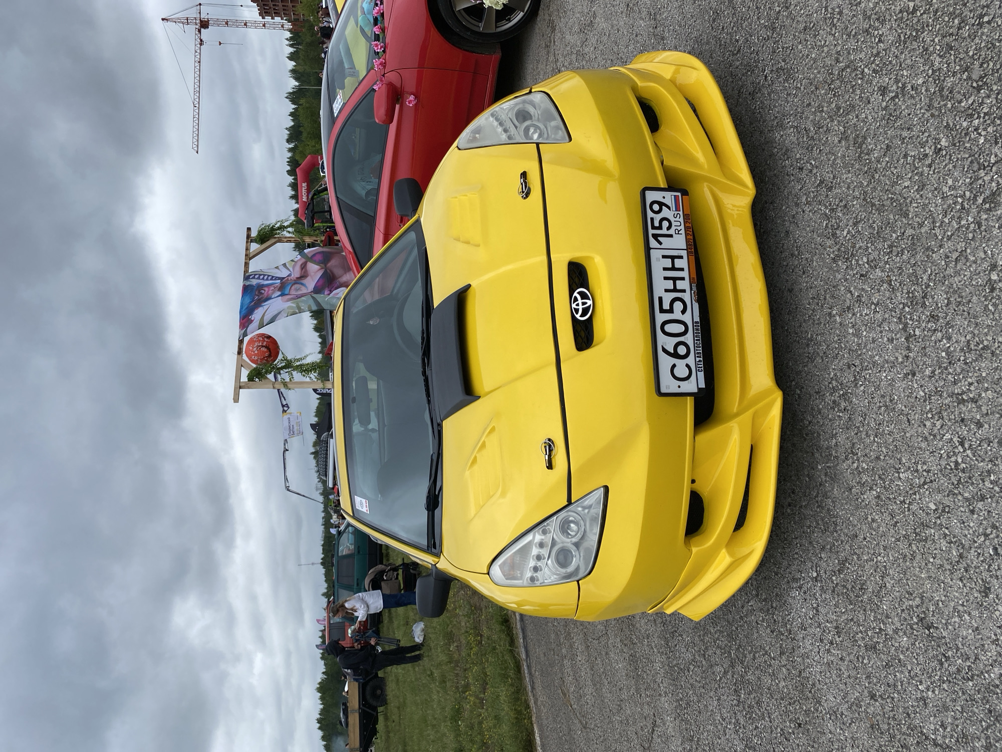 The image depicts an outdoor event, likely a car show, in the city of Perm, Russia. The foreground is dominated by a bright yellow Toyota Celica, featuring a custom body kit and a black hood scoop. Its license plate reads "C605HH 159 RUS." To the right of the yellow car, a red Toyota Celica is partially visible, adorned with small pink flowers. In the background, a wooden archway with a banner featuring anime-style artwork and a red spherical lantern stands prominently. A construction crane and unfinished buildings are visible in the far distance, suggesting a developing area or proximity to ongoing construction. Several people are present, some standing near the cars, while others are gathered in the mid-ground. The overall atmosphere appears casual and festive. The sky is overcast with heavy clouds.