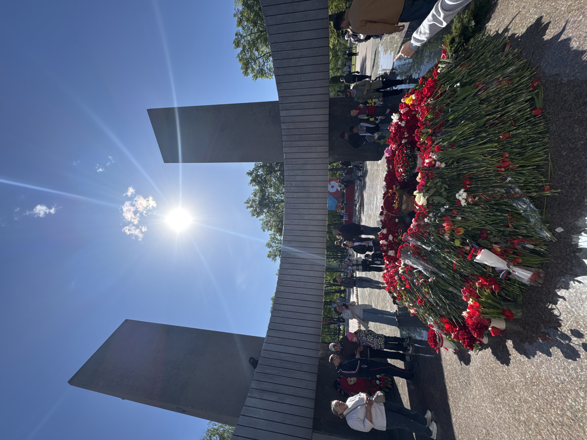 The image shows a memorial with people laying flowers. The memorial is made of concrete and has a large cross-shaped structure. There is a large number of red and white flowers at the base of the memorial. People are standing around the memorial, some are laying flowers. The environment appears sunny, with trees visible in the background. The location is in the city of Unknown, Unknown.