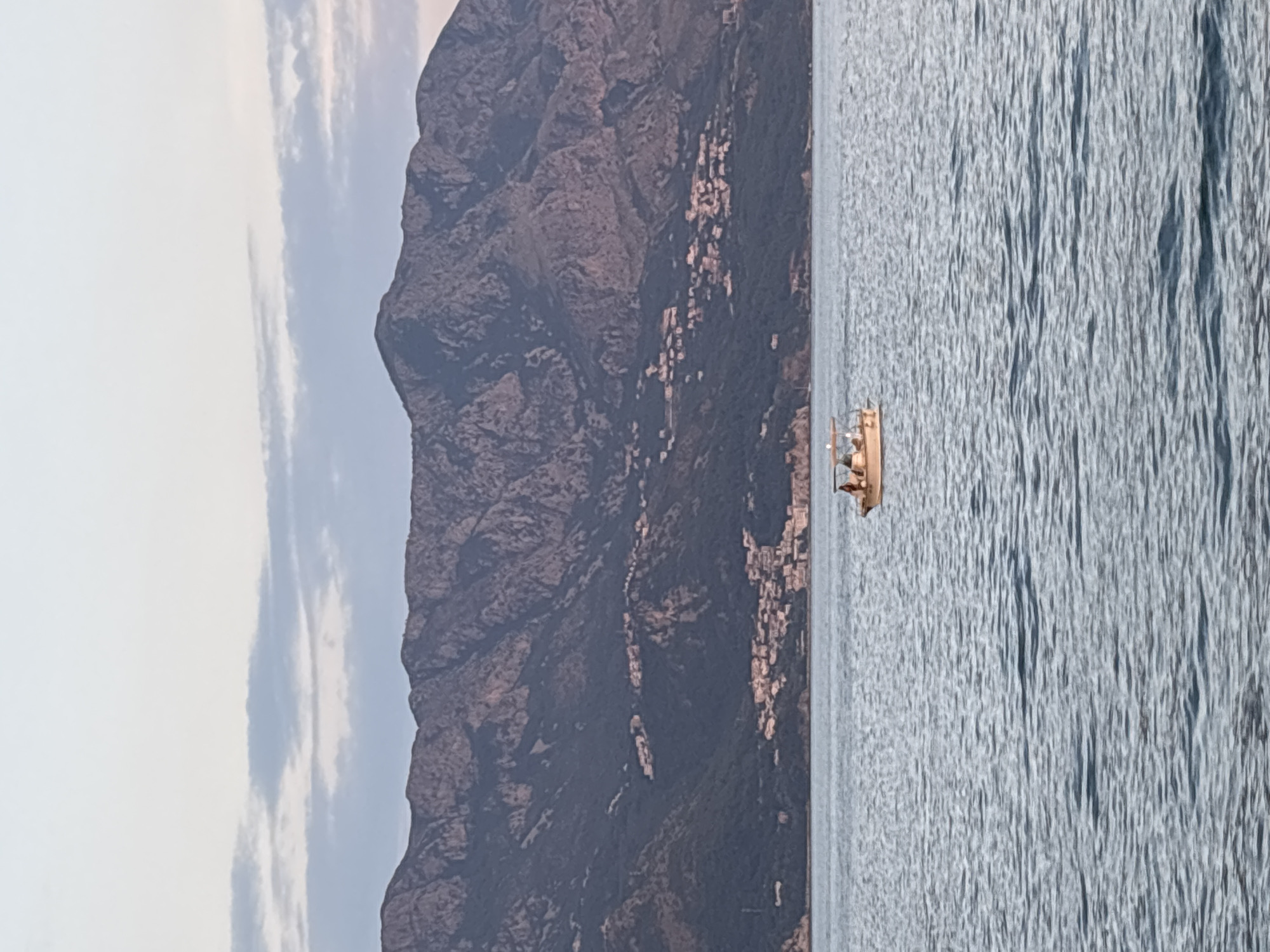 A small motorboat with several people aboard travels across the calm blue waters of the Adriatic Sea. In the background, a range of steep, rugged mountains rises dramatically, dotted with clusters of white buildings that make up the town of Budva, Montenegro. The sky above is a soft gradient of pale blue, with wisps of white clouds.