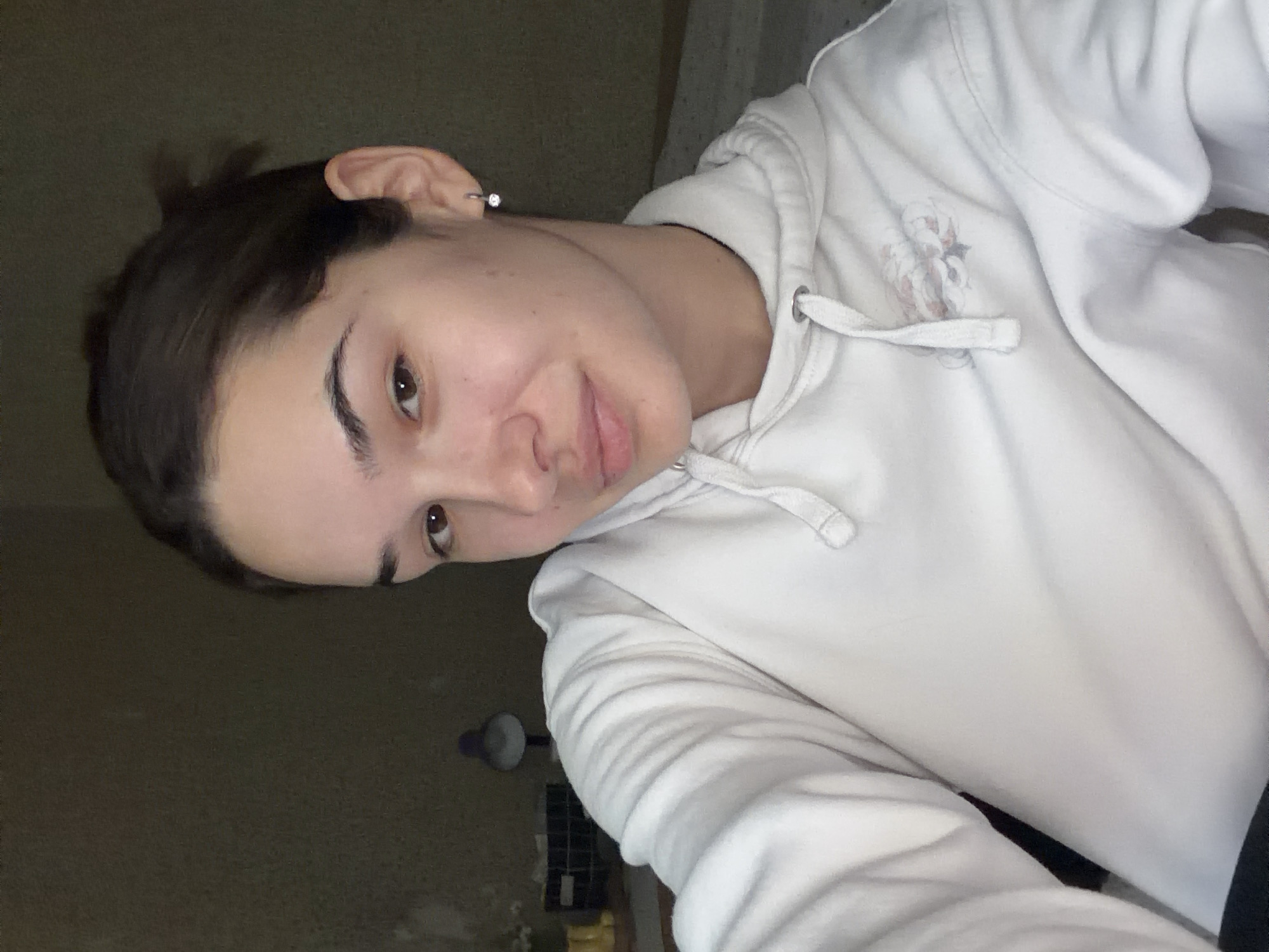 This image is a dimly lit, tilted selfie of a young person, likely female, taken indoors in Sofia, Bulgaria. The subject has dark hair pulled back, dark brown eyes, and fair skin with natural skin variations. They are looking directly at the camera with a gentle, relaxed expression and a subtle, closed-mouth smile.
The person is wearing a white hooded sweatshirt (hoodie) with white drawstrings. A very faint, light-colored graphic design, possibly an abstract or stylized pattern, is subtly visible on the front of the hoodie near the chest. A small, sparkling stud earring is worn on the upper cartilage of the subject's right ear.
The background is largely dark, out of focus, and indistinct, suggesting a dimly lit room, possibly at evening or night. Vague shapes in the background hint at furniture and a distant light source, potentially a lamp. The overall mood conveyed is casual and informal. No visible text is present in the image.