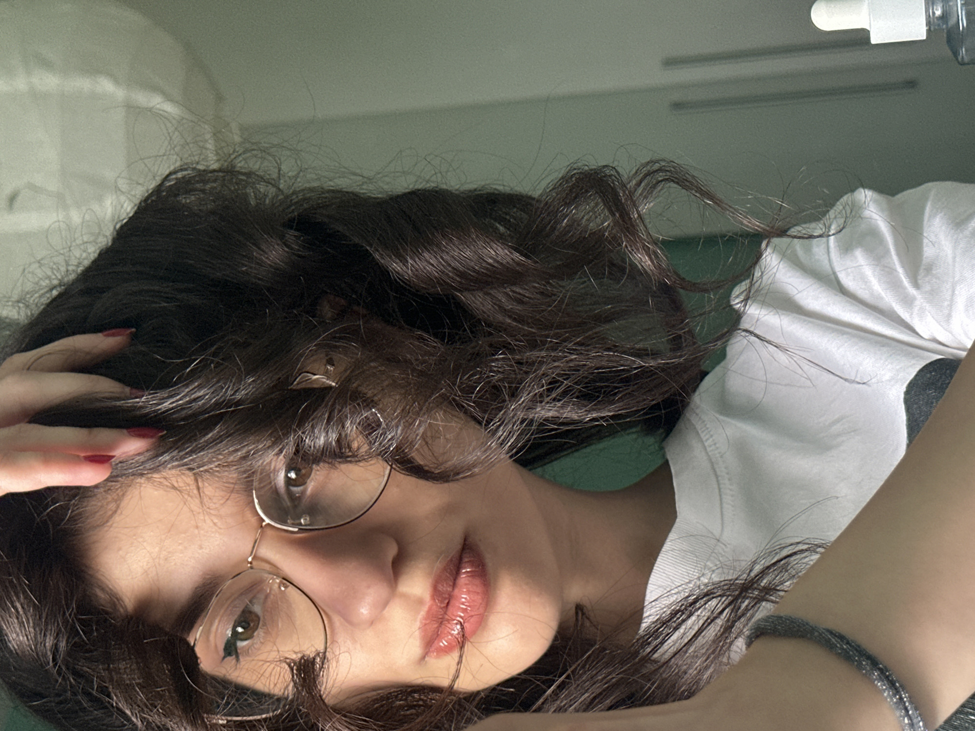 A young woman with dark, curly hair and glasses is shown in a close-up shot, with her hand touching her hair. She is wearing a white t-shirt and a bracelet. Her lips are glossy and her eyes are looking towards the camera. The background is a plain, light green wall with a door and a small bottle with a dropper in the lower right corner. There is no discernible indication of the city of Sofia, Bulgaria in the image.