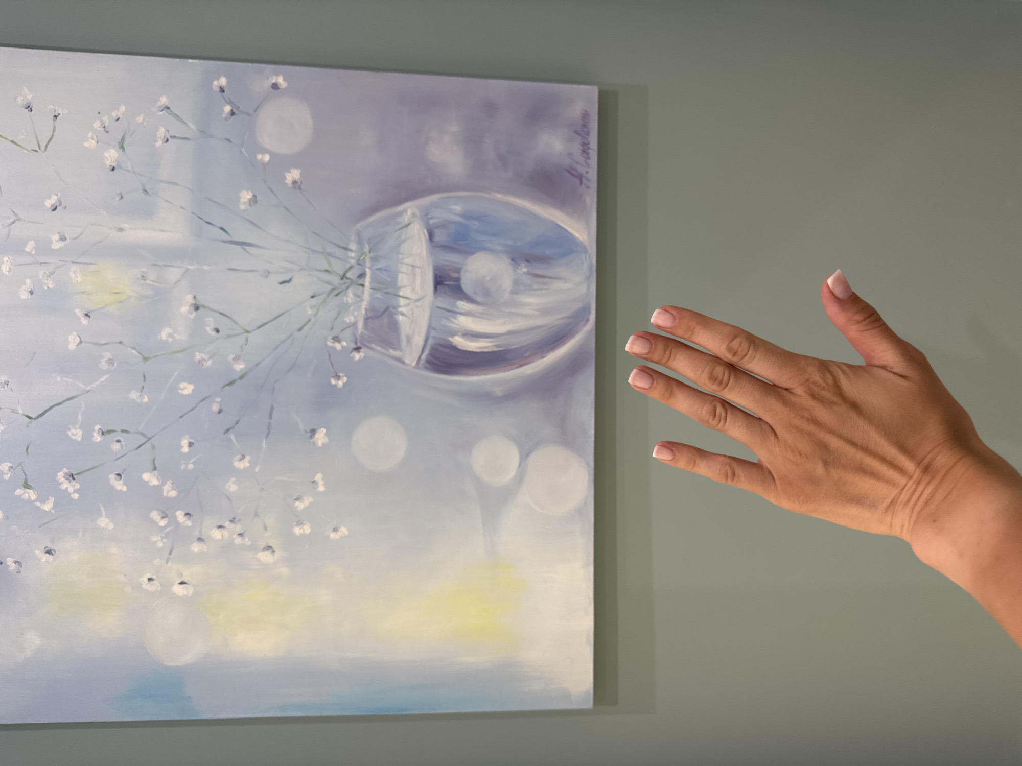 An artist's hand with French manicured nails is held up in front of a painting. The painting depicts a glass vase filled with water and delicate white flowers against a soft, abstract background of blues, whites, and pale yellow. The painting appears to be signed in the lower right corner with a flourish of purple paint, possibly the artist's signature. The background wall is a muted green. The scene is indoors, likely in an art gallery or a residential setting. There are no identifiable landmarks that place this image specifically within Sochi, Russia.