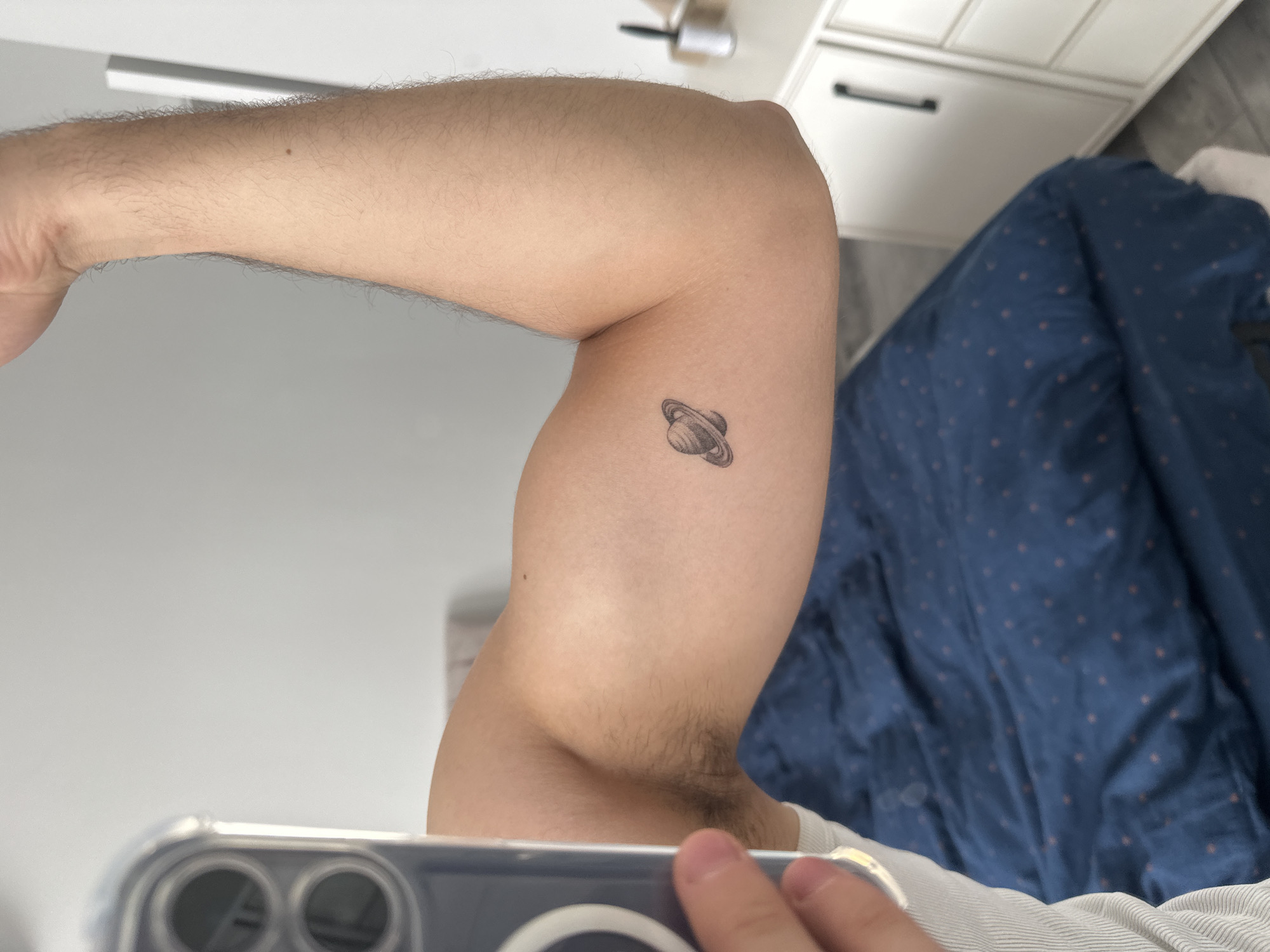 The image shows a close-up of a person's arm, with a small tattoo of Saturn on the bicep. The arm is light-skinned and lightly haired. The person is wearing a white t-shirt. In the background, a blue comforter with a subtle pattern is visible, along with the corner of a dresser with white drawers and black handles. The overall lighting suggests an indoor setting, possibly during the daytime. The focus is on the tattoo and the arm, implying the media is intended to showcase the tattoo. The reflection in the lower part of the image shows a smartphone, suggesting the photo was taken by the person themselves. There is no visible text in the image.