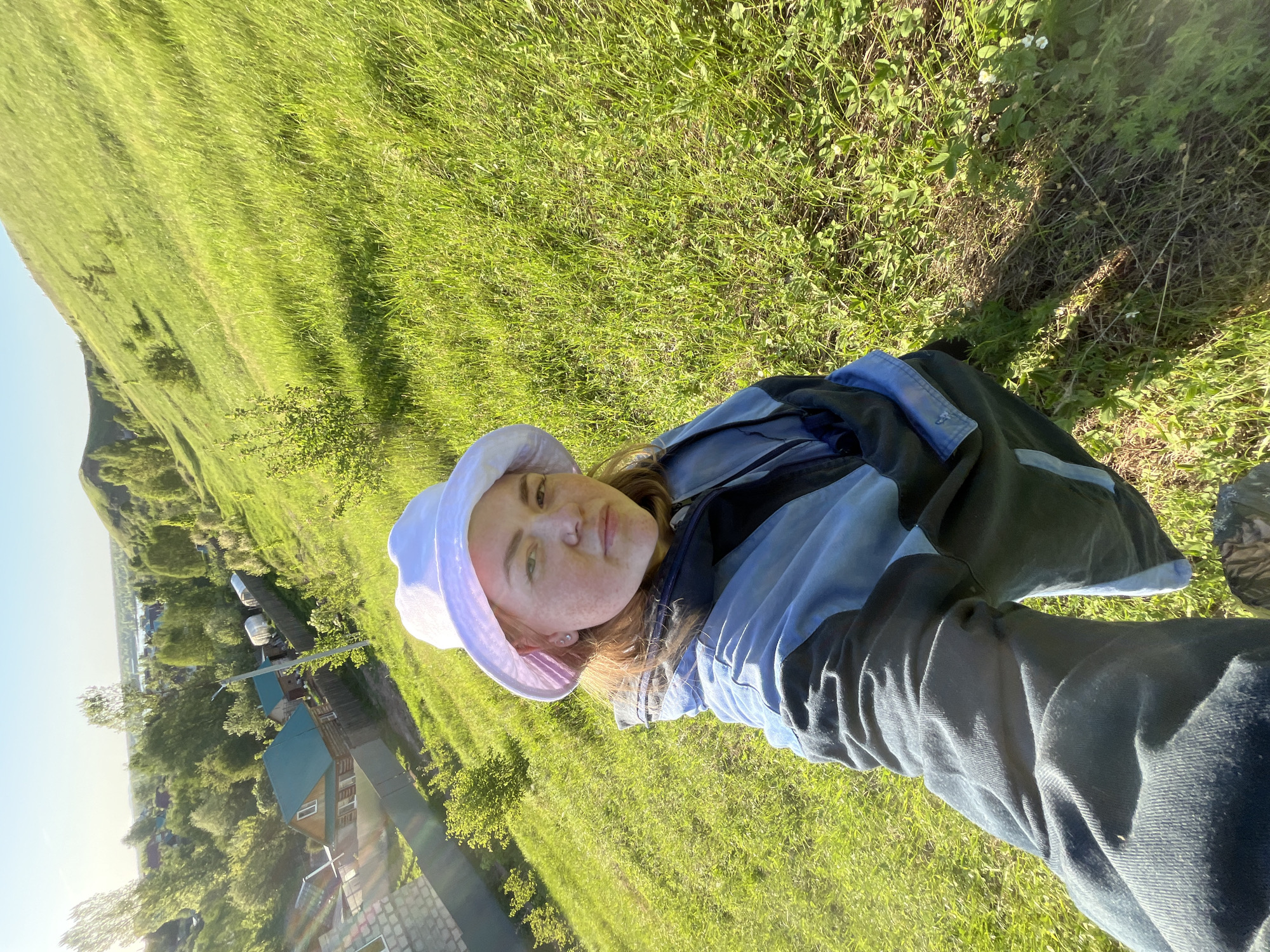 A selfie of a female subject wearing a light-colored bucket hat and a blue jacket is in focus. She is positioned in front of a background of lush green grass. In the distance, there are buildings and structures, likely residential, nestled amidst trees, indicative of a village setting. A hill or low mountain rises in the background. The photo appears to be taken in a rural area within the city of Ольховка, Russia, captured during daylight hours.