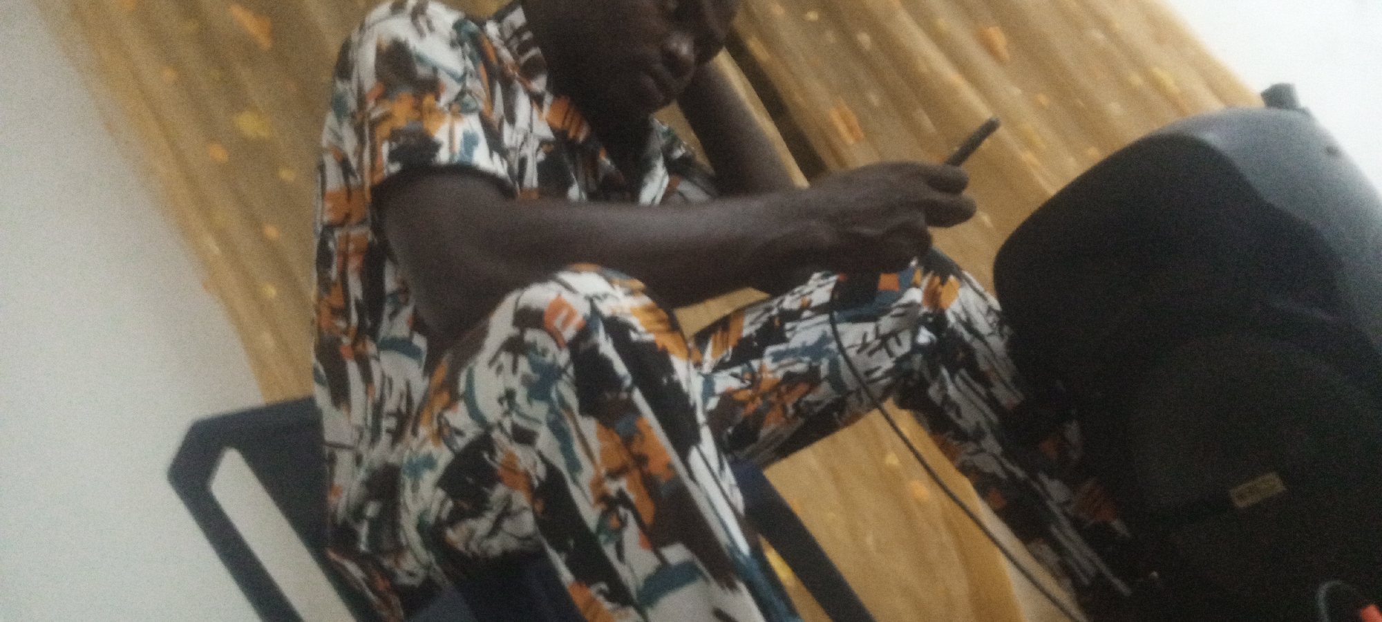 A person in a patterned shirt and pants is sitting on a chair and using a smartphone. To the right is a black speaker. The background features a light-colored curtain. The location is Unknown, Unknown.