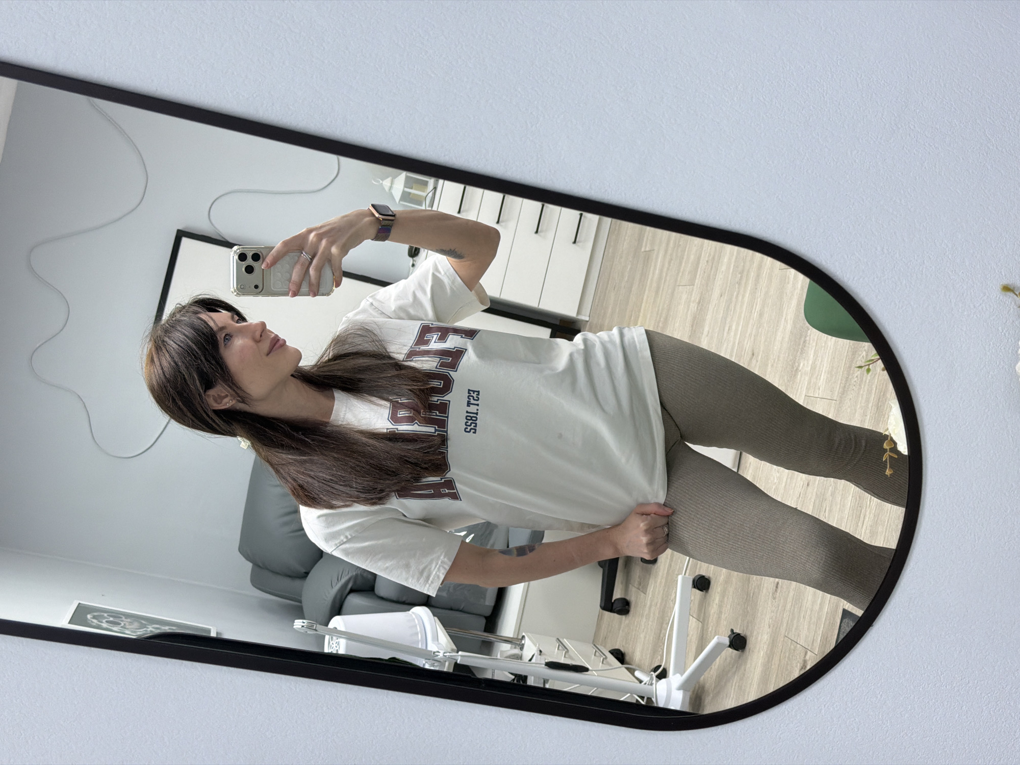 A woman is taking a selfie in a mirror. She is wearing a white t-shirt with a maroon logo and grey leggings. She is holding a smartphone in her right hand, and she is looking at the screen. The mirror has a black frame and an irregular shape. The background of the image includes a grey chair, a white table, and a white dresser. The scene is indoors, and the lighting is bright. The location is likely inside an apartment or home, but the city of Perm, Russia cannot be determined from the image.
