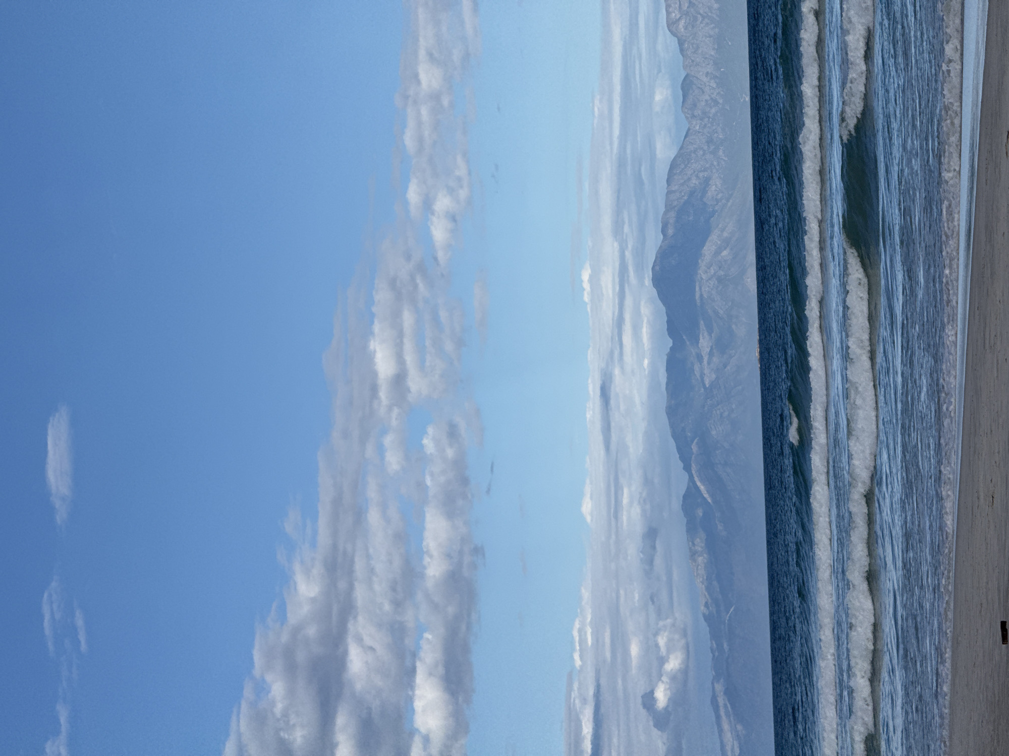 The image captures a beach scene in Cape Town, South Africa. To the left is a bright blue sky with fluffy clouds. Adjacent to the sky are towering, pale, rocky mountains. Next to the mountains is the blue ocean with white waves breaking along the shoreline. Finally, to the right is tan sand.