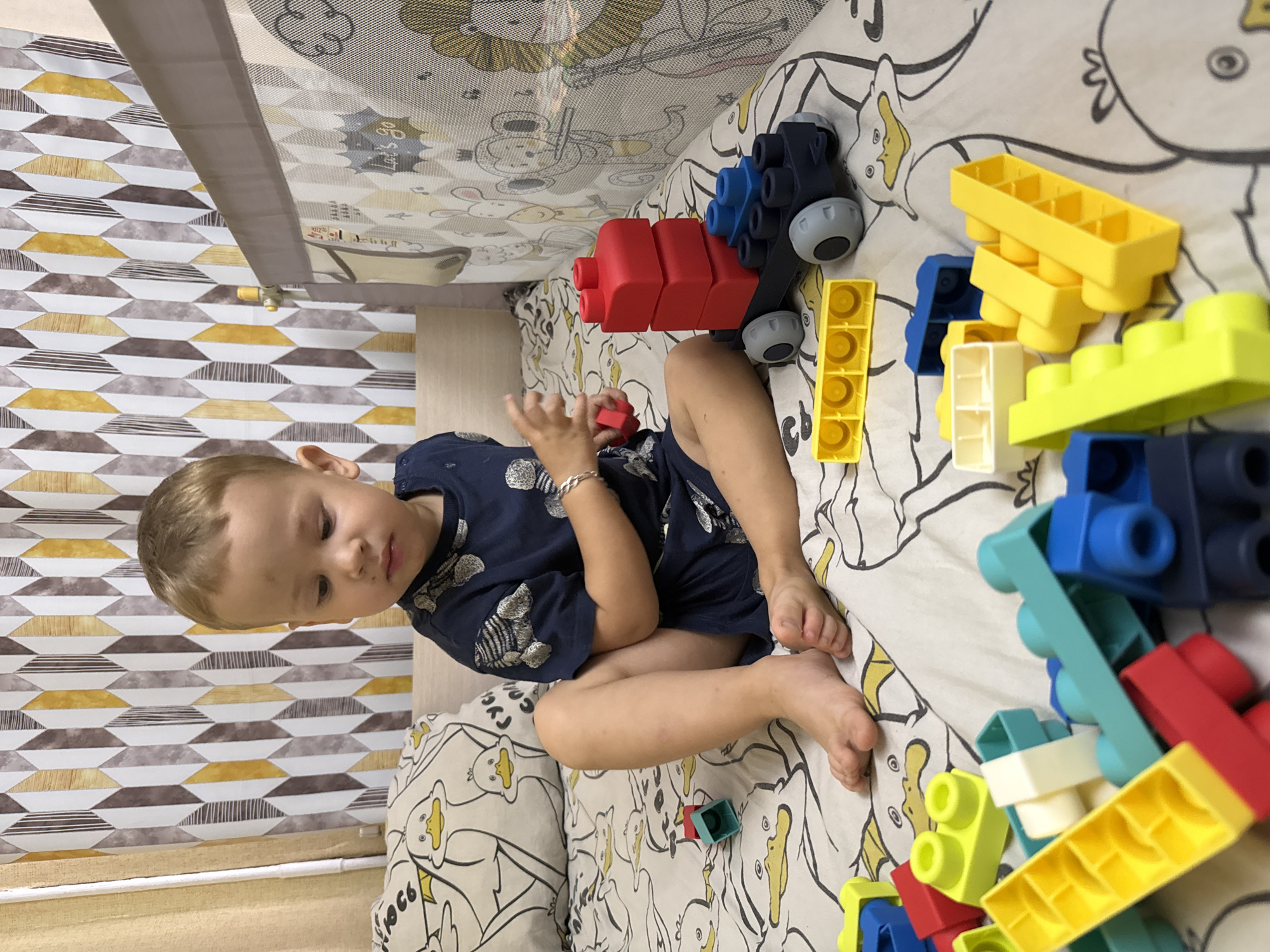 A young boy with blond hair sits on a bed covered with a sheet featuring a cartoon duck pattern. He is wearing a dark blue t-shirt with a printed design and dark blue shorts. The boy is looking down and playing with a collection of colorful building blocks. Scattered around him are various blocks in red, blue, yellow, green, and white, as well as a partially assembled toy vehicle. In the background, a patterned curtain with a geometric design in shades of brown, white, and yellow is visible. To the right, a section of a crib or playpen with a mesh netting and a printed pattern of animals is present. The scene appears to be indoors, likely in a child's bedroom. There is no information in the image to confirm or deny the location as Rusanovo, Russia.