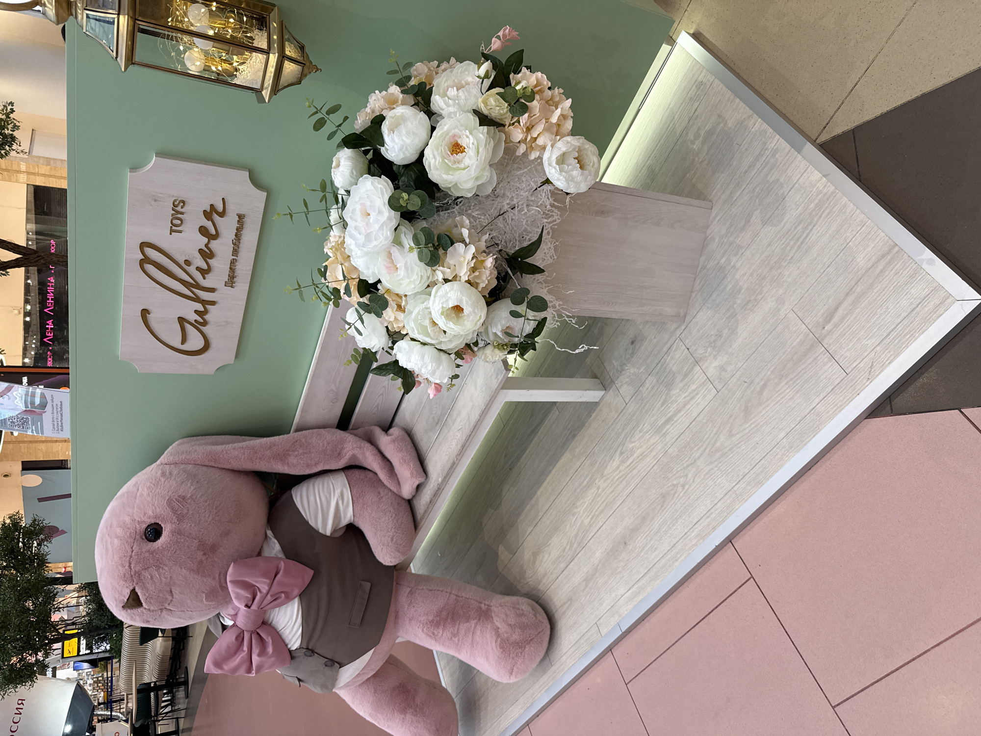 A plush pink bunny toy with a brown vest and pink bowtie sits on a light-colored wooden bench. A bouquet of white and peach-colored flowers is in a light wooden vase to the right of the bench. Behind the bench is a mint-green wall with a light wooden sign that reads "Gulliver TOYS" in gold lettering. A brass lantern with fairy lights is hanging on the right side of the wall. The scene is set on a light wooden floor with a pink tiled floor to the left. The backdrop suggests an indoor retail environment, possibly a department store or mall, given the architectural elements and signage. The overall impression is a decorative display for a toy store, likely "Gulliver Toys," a prominent Russian toy retailer with locations in Moscow.