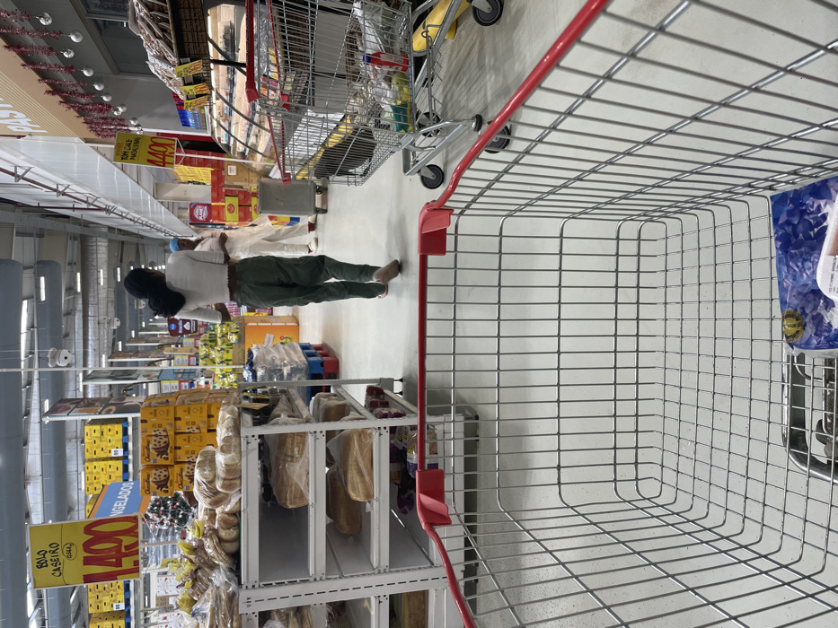 An image captured from the perspective of a shopping cart shows a supermarket aisle. In the foreground, the empty wire basket and red handle of a shopping cart are prominent. A woman with dark hair, wearing a light-colored top and dark green pants, walks away from the camera down the wide aisle. She is in motion, with her right foot slightly lifted. Further down the aisle, two other individuals in light clothing are partially visible.
The supermarket is brightly lit with artificial lighting. On the left side of the aisle, numerous shelves are stocked with various products. There are stacks of boxed goods, possibly cookies or biscuits, and plastic-wrapped bread loaves on lower shelves. A large yellow sign on the left reads "BOLO CASEIRO 1.490" (Homemade Cake 1.490). Below it, another sign partially visible states "CONGELADOS" (Frozen). Further down the aisle, another yellow sign advertises "TOM GALO 1.400" (Gallo Tomato 1.400). The prices, featuring values like 1.490 and 1.400, suggest a local currency. On the right side of the aisle, beyond the foreground cart, more shelves and products can be seen. Another shopping cart, appearing partially loaded, is situated behind the woman. The activity depicted is grocery shopping in a well-stocked and orderly supermarket environment in Luanda, Angola.