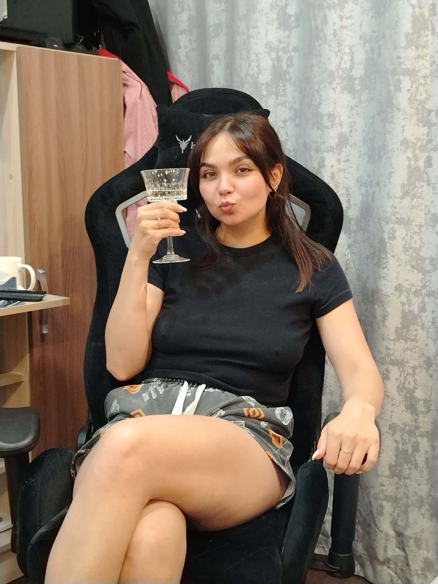 A young woman is captured sitting in a black gaming chair indoors, likely in a room in Noviy, Russia. She is looking directly at the camera with a playful "duck face" expression, puckering her lips. She has brown hair and is wearing a black short-sleeved t-shirt and grey shorts adorned with orange and white patterns, possibly depicting abstract shapes or pumpkins. A simple gold-colored ring is visible on her left ring finger.
In her right hand, she holds up a decorative glass containing a light-colored, effervescent beverage, possibly sparkling wine or champagne. The black gaming chair has a distinctive white, stylized logo on its headrest, resembling a bird or abstract symbol. To the woman's left, part of a wooden cabinet or wardrobe is visible, with a red and white striped shirt and a black garment draped over it. A small, floating shelf attached to the cabinet holds a white mug with a dark design and another dark rectangular object. The background wall on the right features a light grey patterned wallpaper. The scene conveys a relaxed and casual moment.