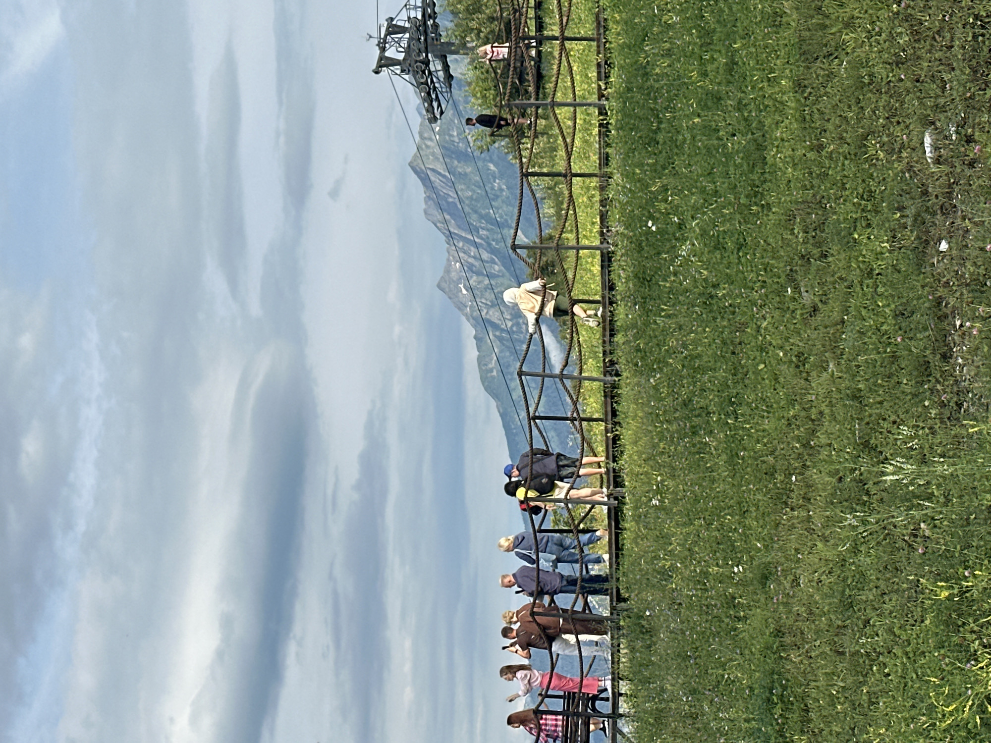 The image depicts a scenic mountain landscape in Estosadok, Russia, likely during the daytime under a cloudy sky. A wooden walkway, supported by metal posts and a rope railing, stretches horizontally across the frame, offering a viewing platform. A group of people are standing on the walkway, some taking pictures with their phones, others observing the scenery. In the background, a ski lift mechanism is visible, with its cables extending towards the mountains. The mountains themselves are covered in a mix of green vegetation and rocky terrain, with some snow patches on their peaks. A dense field of green grass, dotted with small white and pink flowers, occupies the foreground. The overall atmosphere suggests a popular tourist destination, with people enjoying the natural beauty of the mountainous region.