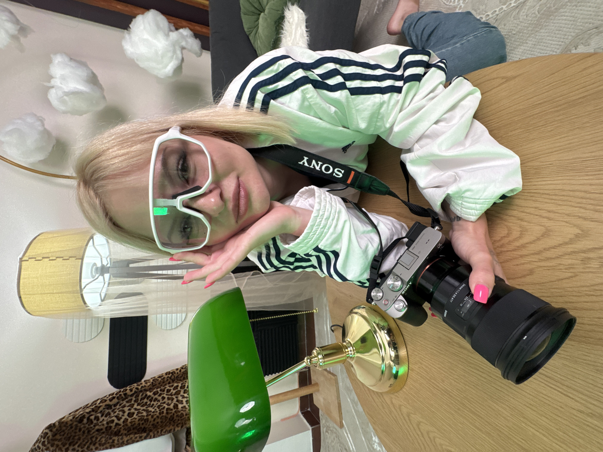 A fair-skinned female, approximately 20-30 years old, with blonde hair styled with bangs and shoulder-length layers, is positioned on the floor, leaning on a light-colored wooden table in an indoor setting in Budva, Montenegro. She is wearing white, wide-framed protective or sports-style glasses with tinted lenses and a small green rectangular accent on the left lens. Her nails are painted bright pink. She is dressed in a white Adidas track jacket with three black stripes extending down each sleeve.
Her left hand, resting on her cheek with her elbow on the table, supports her head as she looks directly at the viewer with a slight pout. Her right hand firmly grasps a silver and black Sony mirrorless camera, identifiable by the "SONY" branding on its strap and body, fitted with a large black Sigma 24mm 1:1.4 DG DN Ø77 lens. A nose piercing is visible on her left nostril.
To her left, on the wooden table, is a vintage-style green banker's lamp with a polished brass base and stem. The background reveals an eclectic decor. On the left, a brown and black leopard print fabric is draped, and a wall features light-colored circular or oblong decorative panels and a horizontal black panel. A tall floor lamp with a yellow-tan fabric shade is partially visible behind these elements. On the right, several fluffy white cloud-like decorations are affixed to a white wall. Below these, a dark gray sofa with a round green cushion and a white furry cushion is visible. The floor is covered with a light-colored patterned rug.