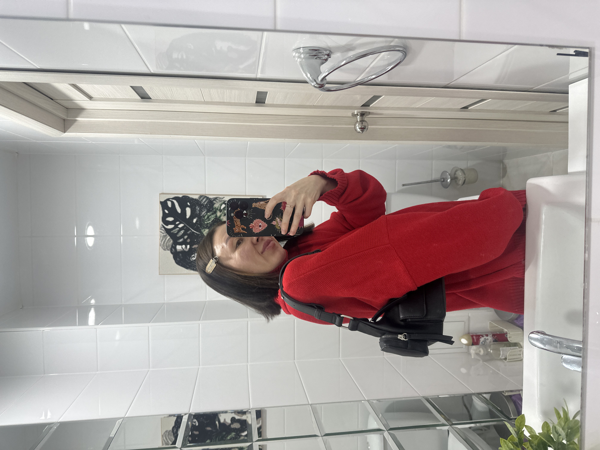An interior mirror selfie taken in a bathroom in Votkinsk, Russia, depicts a female subject. The individual, dressed in a red long-sleeved sweater and a black shoulder bag, faces slightly left while holding a smartphone with a patterned case (animal print and stylized hearts/eyes) to capture her reflection. A ring is visible on her left ring finger. Her dark, shoulder-length hair is accessorized with a light-colored hair clip on the right side. The bathroom features white tiled walls and a white ceramic sink with a chrome faucet in the foreground. A light-colored wooden door with a chrome handle and a dark rectangular insert is visible to the right of the subject's reflection. A chrome ring-shaped towel holder is mounted on the wall to the right of the door. Reflections reveal a framed botanical artwork with a black and white leaf pattern, a small white basket containing a red spray can and a clear pump bottle behind the sink, a toilet brush in its holder near the floor, and a small green potted plant near the left edge of the sink. A wall section composed of angled mirror panels or mirrored tiles is visible on the far left.