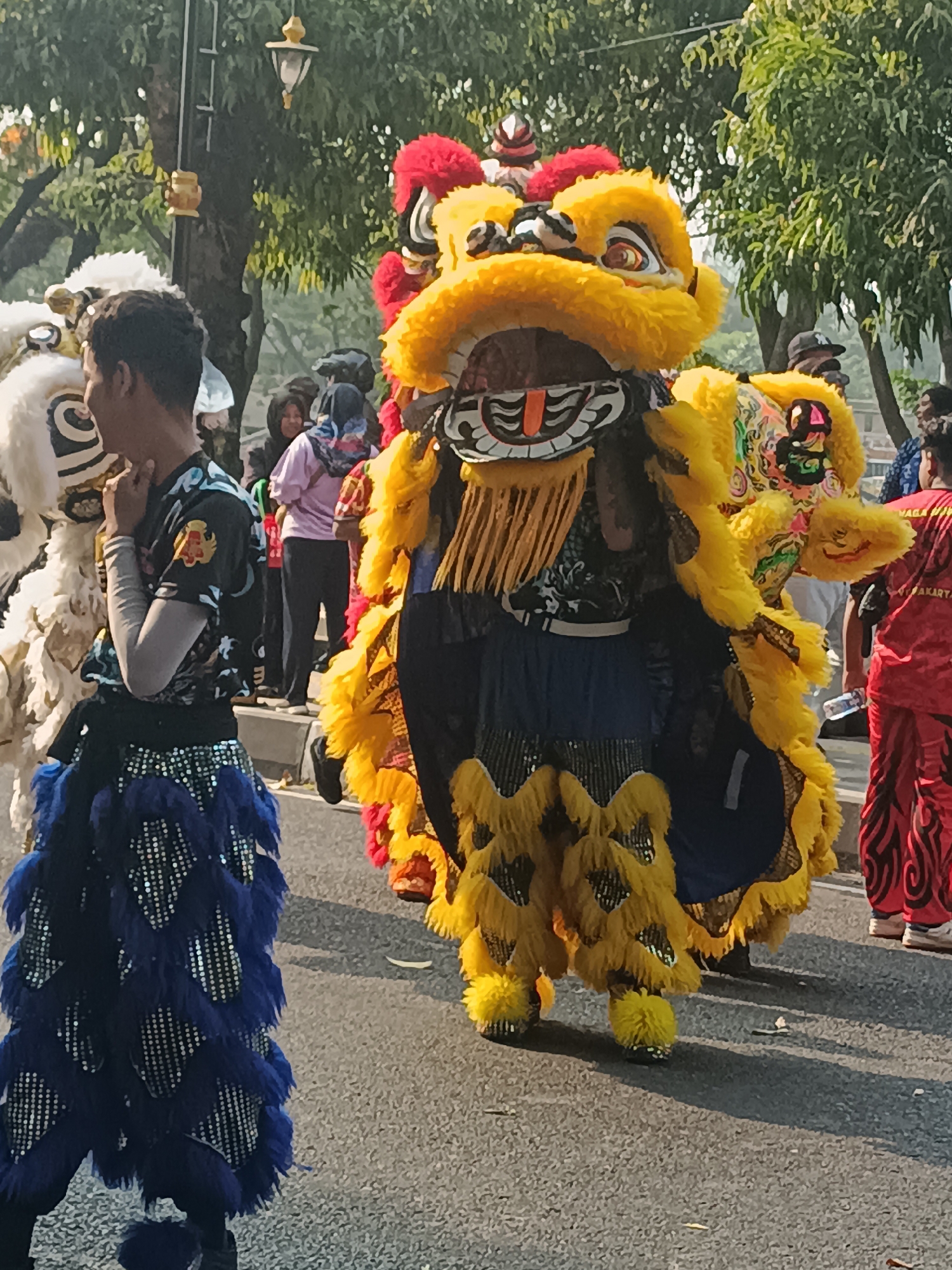 The image depicts a street scene in Sleman, Indonesia, likely during a cultural event or parade. In the foreground, a person in a vibrant yellow and blue lion dance costume is prominently featured. The costume is elaborate, with a large, furry yellow head adorned with red and black accents, and a blue skirt-like lower body covered in sparkling sequins. To the left, another lion dance costume, this one predominantly white and black with blue accents, is partially visible, with a person inside wearing a dark t-shirt with a colorful emblem. Behind the main subjects, several other individuals are present, some wearing traditional attire, including a woman in a pink headscarf and another in a red outfit with black patterns. The background consists of trees and a street, indicating an outdoor urban environment. The lighting suggests it is daytime, with shadows cast on the asphalt road.