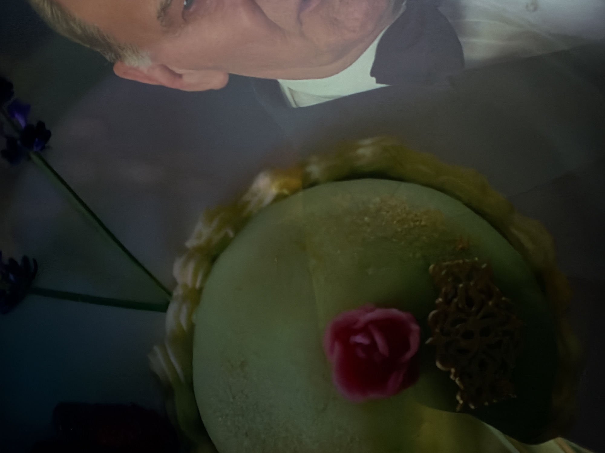 An inverted reflection of a man's face and upper torso is visible in the upper part of the image. The man appears to be older with light hair, fair skin, and is wearing a white collared shirt beneath a dark suit or jacket. His eye and ear are discernible as he looks downwards.
In the foreground, taking up the majority of the lower image, is a light green, round object, possibly a cake or a decorative hat, with a textured surface. Adorning this object is a small, vibrant pink decoration shaped like a rose, positioned towards the left. To its right, there is an intricate, gold-colored filigree or lace-like embellishment.
To the left side of the frame, several dark purple flowers with slender green stems are visible, hinting at a floral arrangement. The overall lighting is dim, suggesting an indoor setting, possibly during the evening or under artificial light. The blurred quality of the reflection contrasts with the clearer focus on the green object. No text is visible in the image. The scene suggests a celebratory or formal event. While the location is Sofiivska Borshchahivka, Ukraine, no specific geographical cues are present within the image itself.
