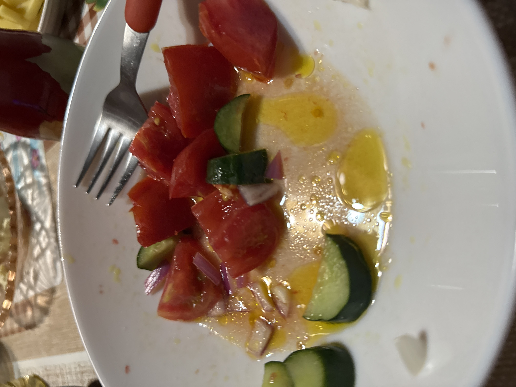 A close-up, high-angle shot shows a partially eaten salad on a white plate. The salad consists of chopped tomatoes, cucumber slices, and red onion. The ingredients are coated in an oil-based dressing that has pooled in the bottom of the plate. A fork with a red handle rests on the edge of the plate, with its tines partially submerged in the salad. The background is blurred but appears to be a dining table with other dishes and a drink bottle. The scene is likely indoors, possibly in a restaurant or home setting in Sarnevo, Bulgaria.