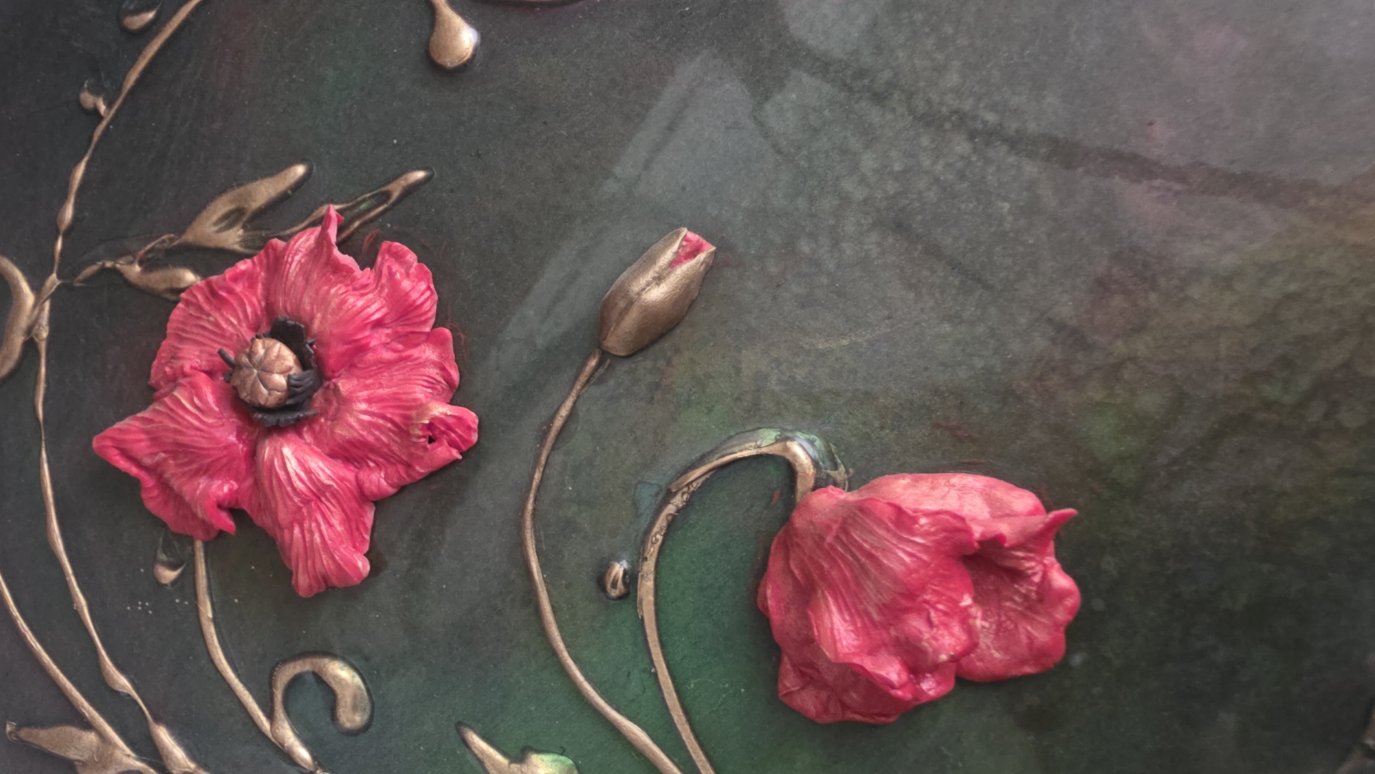 The image shows a close-up of a dark green surface with three-dimensional poppy-like floral designs. Two are fully bloomed, and one is a bud. The flowers are red with dark centers. The stems and accents are bronze-colored and appear to be painted or applied in a relief style. The background has a glossy finish. The image does not provide enough information to determine the location within Saint Petersburg, Russia, or any information about people, interactions, or actions.