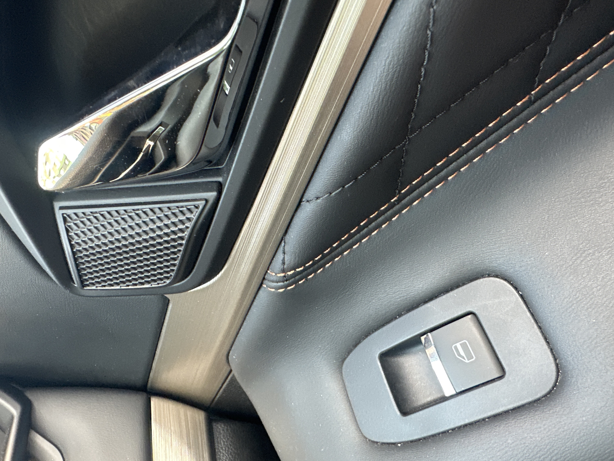 The image shows the interior of a car door, focusing on a window control switch and part of the door handle. The switch is black and rectangular with a silver and white icon depicting a car window. The door panel is upholstered in black leather with prominent orange stitching in a diamond pattern. Above the leather is a metallic trim piece with a brushed finish, followed by a textured black speaker grille and a chrome door handle. The scene is forensic in nature, providing a detailed look at the materials and components of the car's interior. There are no people, actions, or interactions depicted, nor any elements that would suggest a specific location in the city of Ruzino, Russia.