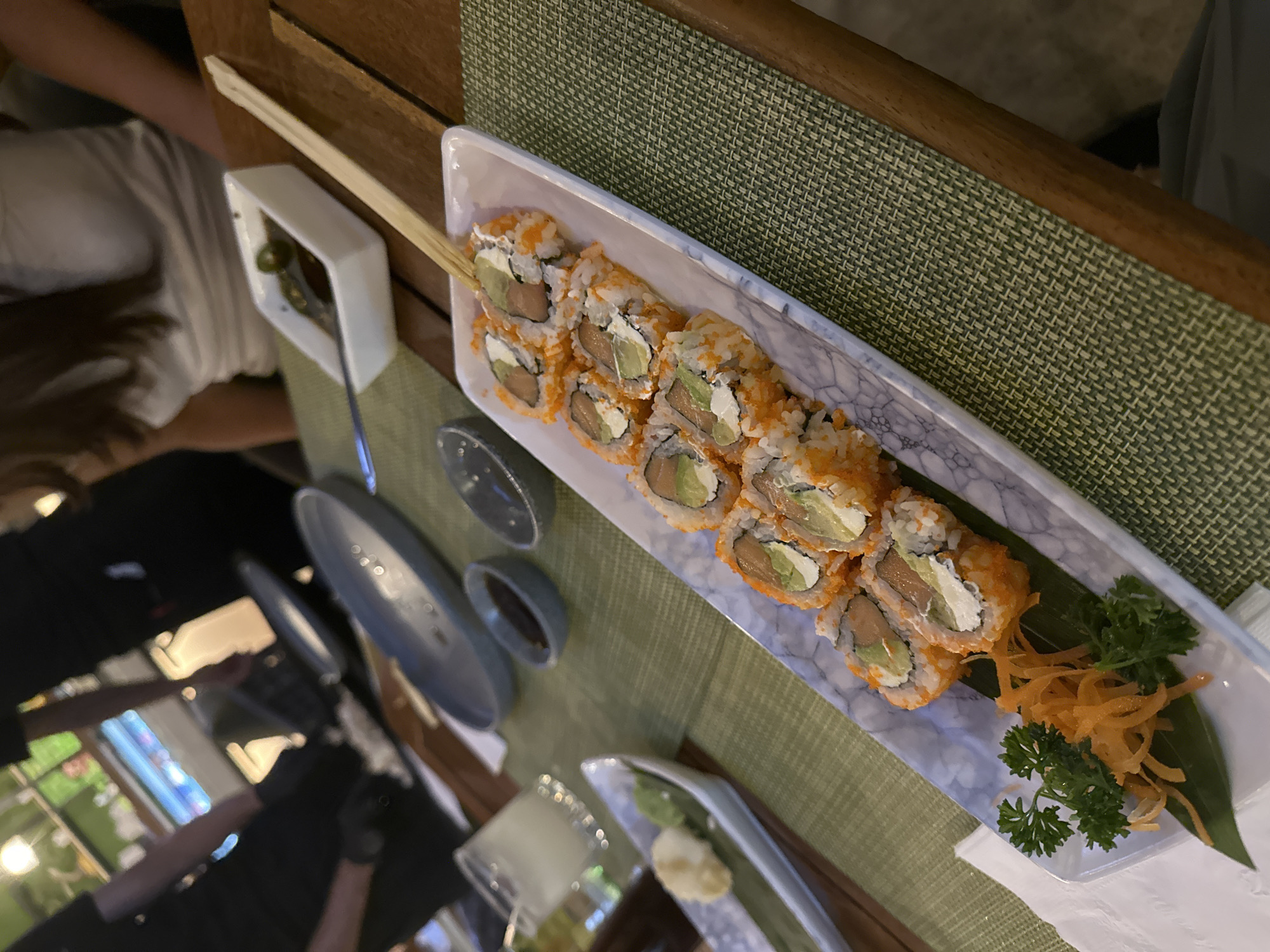 A rectangular white plate of sushi with avocado, salmon, and tobiko topping is on a green woven placemat. A pair of wooden chopsticks rests diagonally across the top of the plate. Several small blue-gray dipping bowls are next to the plate. A small dish containing pickled ginger and wasabi is in the foreground. A partially visible person wearing black gloves is in the lower-left corner. Another person in a white shirt is visible in the upper-left corner. The location is San Bernardino Tlaxcalancingo, Mexico.