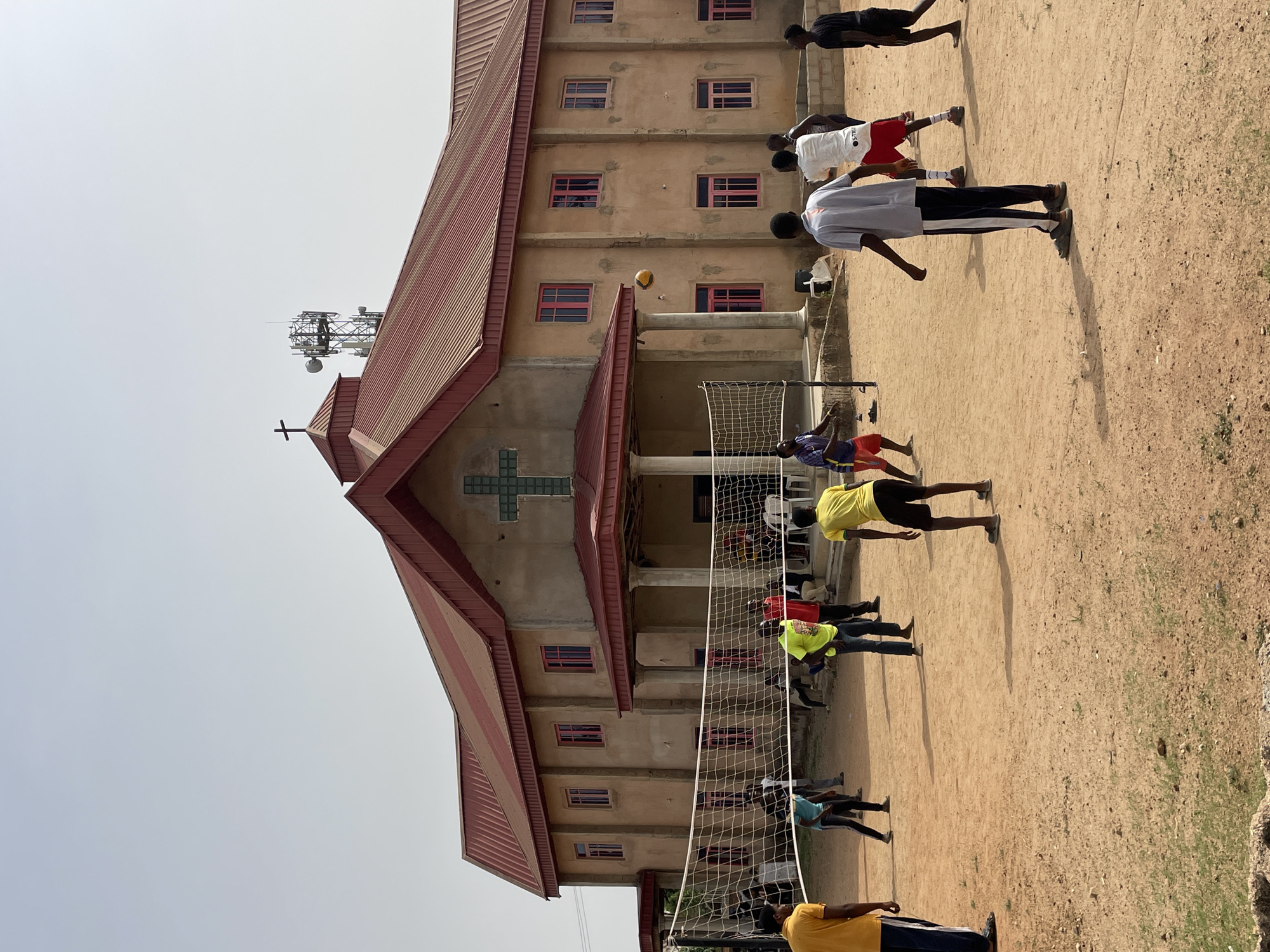 A medium shot captures a group of individuals playing volleyball on a dirt court adjacent to a building. The building is tan with a red roof and features a prominent cross on its facade. A volleyball is mid-air during play. An antenna is atop the building. The scene is likely within the city of Jos, Nigeria.