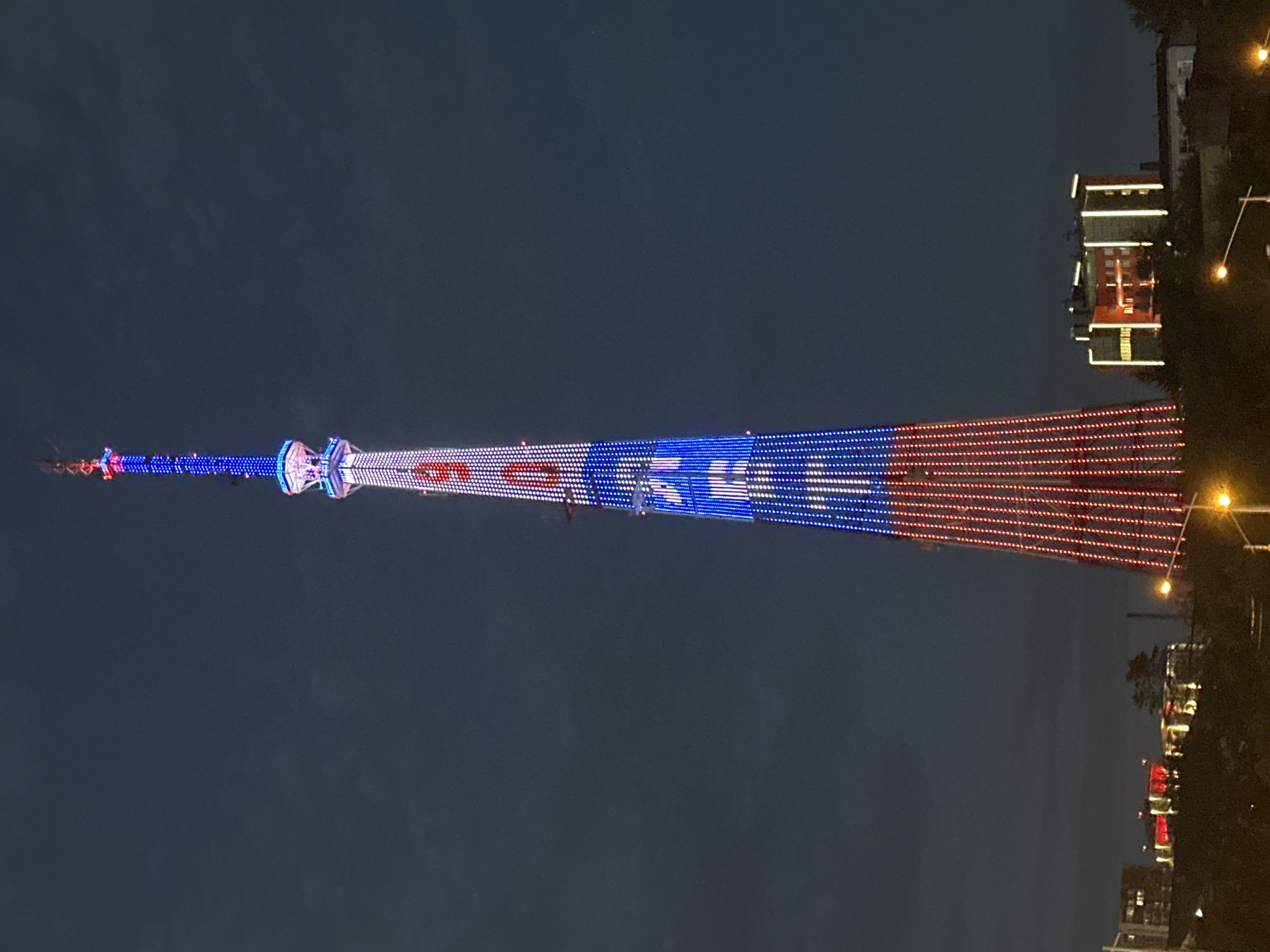 The image captures a nighttime view of a tall tower illuminated with red, white, and blue lights, resembling the colors of the Russian flag. The tower is the main subject, extending diagonally across the frame against a dark, twilight sky. The lights create a vibrant, patriotic display, with horizontal bands of blue LEDs at the top, transitioning to white and then red LEDs towards the bottom. Small clusters of red and blue lights are visible at the very top of the tower, suggesting intricate lighting patterns.
In the background, to the right of the tower, are buildings with illuminated windows and exteriors, indicating an urban setting. These buildings are also lit with warm, yellow lights. A few trees are visible, their dark silhouettes contrasting with the lit structures. Several streetlights with a warm glow are also present, adding to the nighttime ambiance. The overall scene suggests a celebration or a special event taking place, likely in a public area given the scale of the illuminated tower and the presence of buildings and streetlights. The time of day is clearly night or late dusk. No people are directly visible in the image, but the illuminated structures suggest human activity and a populated environment. The location is identified as Irkutsk, Russia, which aligns with the observed patriotic colors of the lighting display.