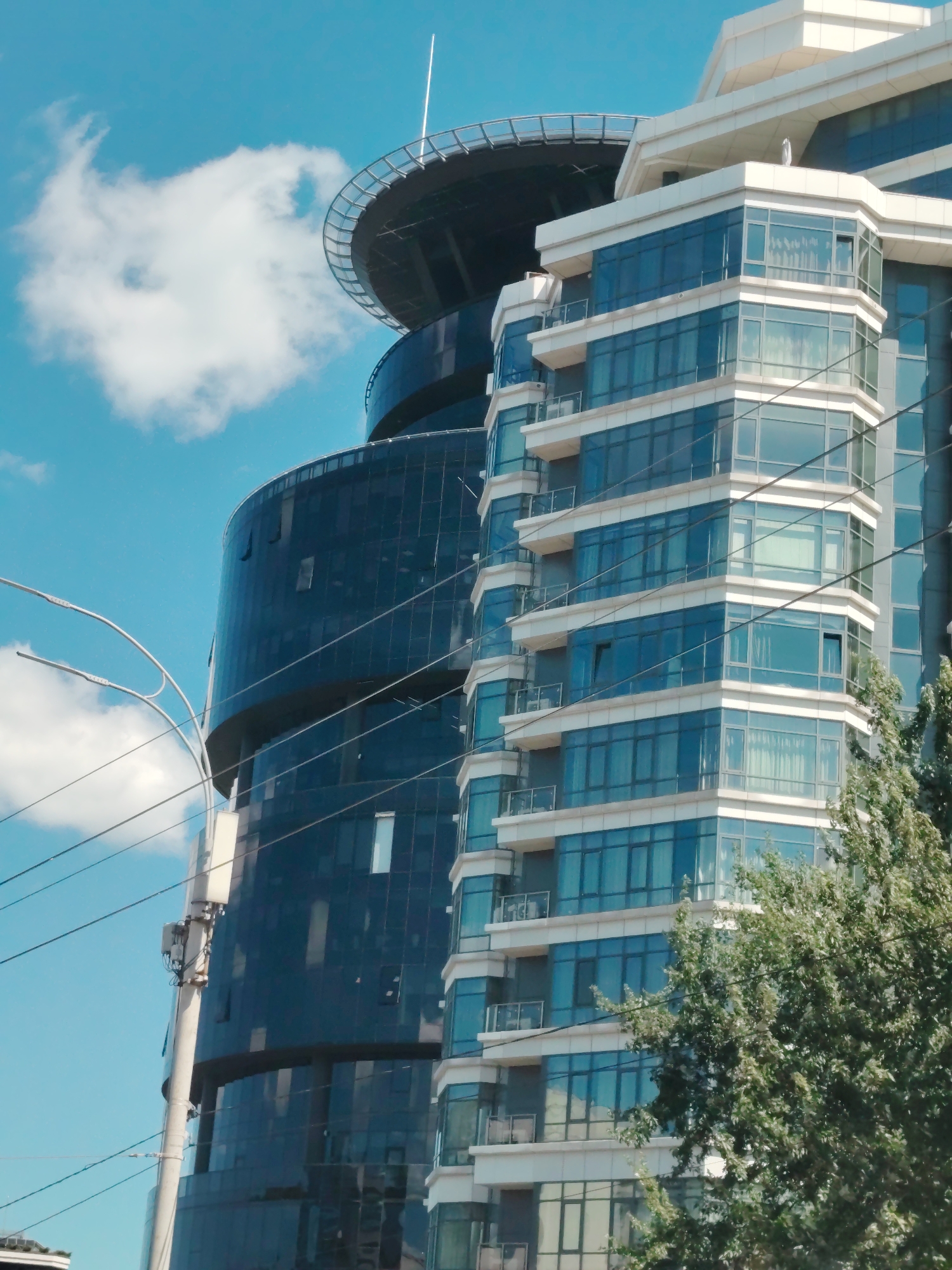 This image depicts a modern architectural scene in Kyiv, Ukraine, focusing on two distinct buildings. On the right, a multi-story building with a white facade and numerous blue-tinted glass windows is prominently featured. The windows are arranged in a pattern across the building's angled surfaces, with balconies visible on some levels. To the left and behind this building is a taller, darker, curved structure with a glass facade. This building has a wide, circular platform at its top, which appears to be a helipad. The sky above is blue with scattered white clouds. In the foreground, a utility pole with wires is visible, and a portion of a green tree obscures the lower right corner of the image. The overall impression is of a contemporary urban environment with a focus on modern architecture.