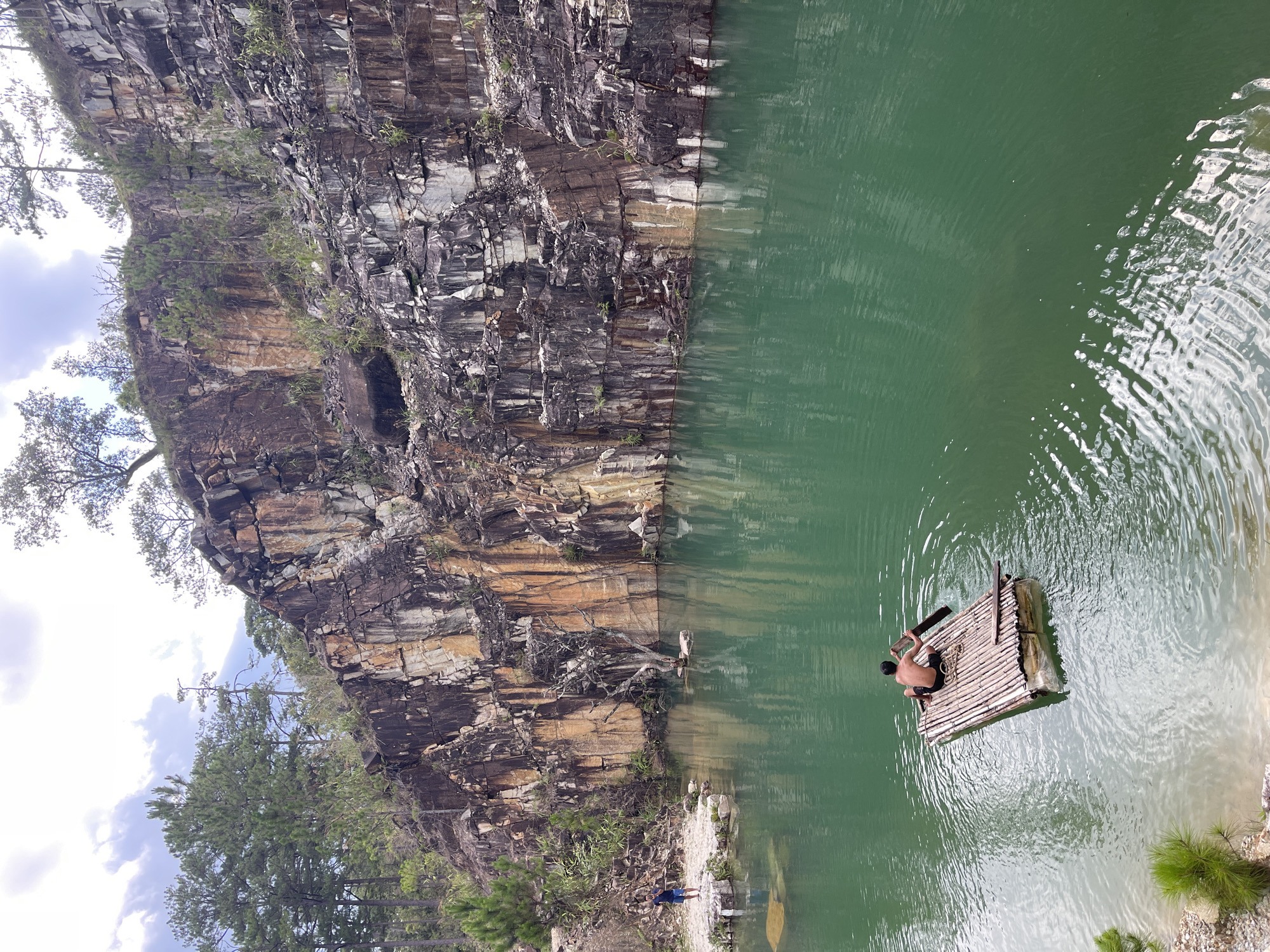 The image depicts a quarry filled with green water in Lang Biang - Da Lat, Vietnam. A rocky cliff forms one side of the quarry. A man is on a bamboo raft in the water, holding a paddle. Another person is standing on the shore. Trees are visible along the top of the cliff and on the surrounding land.