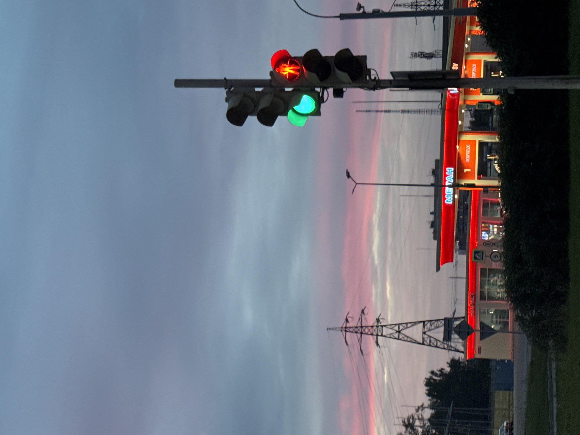 The image depicts a street scene at dusk in Perm, Russia, near a gas station that appears to be a METAN brand. The sky is a gradient of blue and pink hues, suggesting either sunrise or sunset. In the foreground, a traffic light is visible, with the green light illuminated and the red light also glowing, the latter with a pedestrian silhouette. To the left, a large electrical pylon stands against the sky, with power lines extending across the frame. The gas station features bright red signage with "METAH" prominently displayed in blue lettering. Other signs and lights are also visible on the station, including a number "1" and a small blue box next to the station. In the background, there are some trees and other structures, including what appear to be communication towers. A dark green grassy area is present in the immediate foreground. No people are clearly discernible in the image.