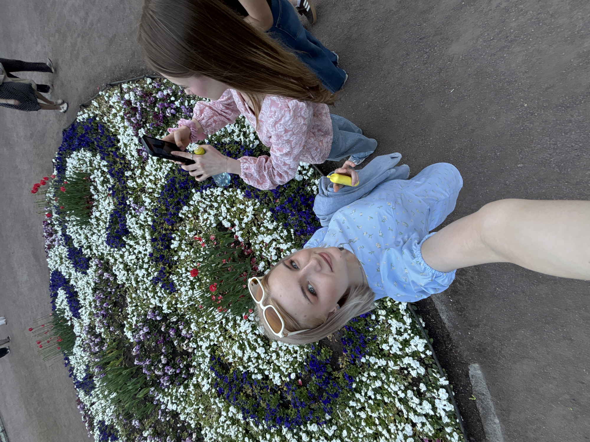 Two young women are depicted standing on a dark, granular path adjacent to a large, elaborately planted flower bed in Saint Petersburg, Russia. The perspective is a wide-angle, high-angle shot, likely a selfie.
The woman in the foreground, positioned to the left, is light-skinned with blonde hair and sunglasses perched atop her head. She is wearing a light blue dress with a subtle floral pattern and looks directly into the camera with a slight smile, holding what appears to be a yellow, cylindrical object in her left hand. Her right arm is extended, indicating she is holding the camera device.
To her right and slightly behind, a second light-skinned woman with long brown hair is visible. She is wearing a pink textured long-sleeved top and light-colored jeans. She holds a black smartphone with both hands, pointed towards the flower bed, appearing to photograph it. A clear plastic water bottle is in her left hand, with another yellow, cylindrical object attached or held near it. Her profile is visible as she focuses on the scene in front of her.
The expansive flower bed features dense plantings of small flowers, primarily white and dark purple/blue, arranged in distinct patterns. Red tulips emerge from green foliage, particularly in the upper and central-right portions of the bed. The edge of the flower bed is dark. In the distant background, several blurred figures walk on the same dark path, suggesting a public park or garden setting. The lighting is soft and even, indicating daytime conditions without direct sunlight.
