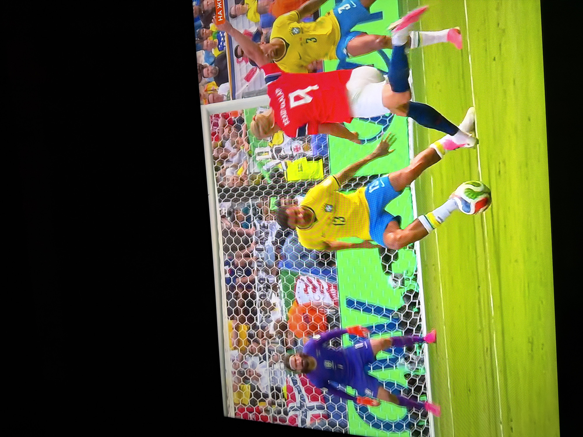 The image depicts a soccer match in progress. Four male athletes are visible on a green field with a soccer goal net in the background. One player in a purple uniform with orange gloves and accents is positioned as a goalkeeper. Two players in yellow and blue uniforms are actively engaged in play, one of whom is dribbling a soccer ball. A fourth player, wearing a red jersey with the number 9, is also on the field. The spectators in the background are blurry and appear to be seated in a stadium. A sign with Cyrillic text, "НА ЖИ", is partially visible in the upper right portion of the image, suggesting the match is being broadcast live. There is no discernible information within the image that places the scene in the city of Ognianovo, Bulgaria.