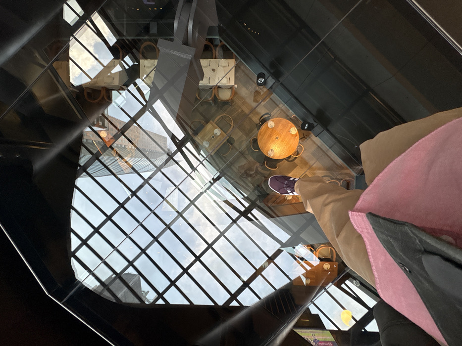 An overhead view captures a person's lower body standing on a transparent glass floor within a building in Mexico City, Mexico. The individual is wearing tan pants and a visible purple athletic shoe with white stripes. A pink bag with a black strap is positioned in the immediate foreground. Below the glass floor, a cafe or restaurant area is visible, featuring a round wooden table set with two clear glasses and surrounded by two light-colored chairs. Additional cafe seating is present further back. The underlying floor is dark and reflective. The transparent surface reflects a grid-patterned skylight showing a cloudy sky, overlaid with the reflection of a tall, modern multi-story building exhibiting a distinct grid facade and sections with a brownish-orange hue. Geometric architectural structures are also reflected.
